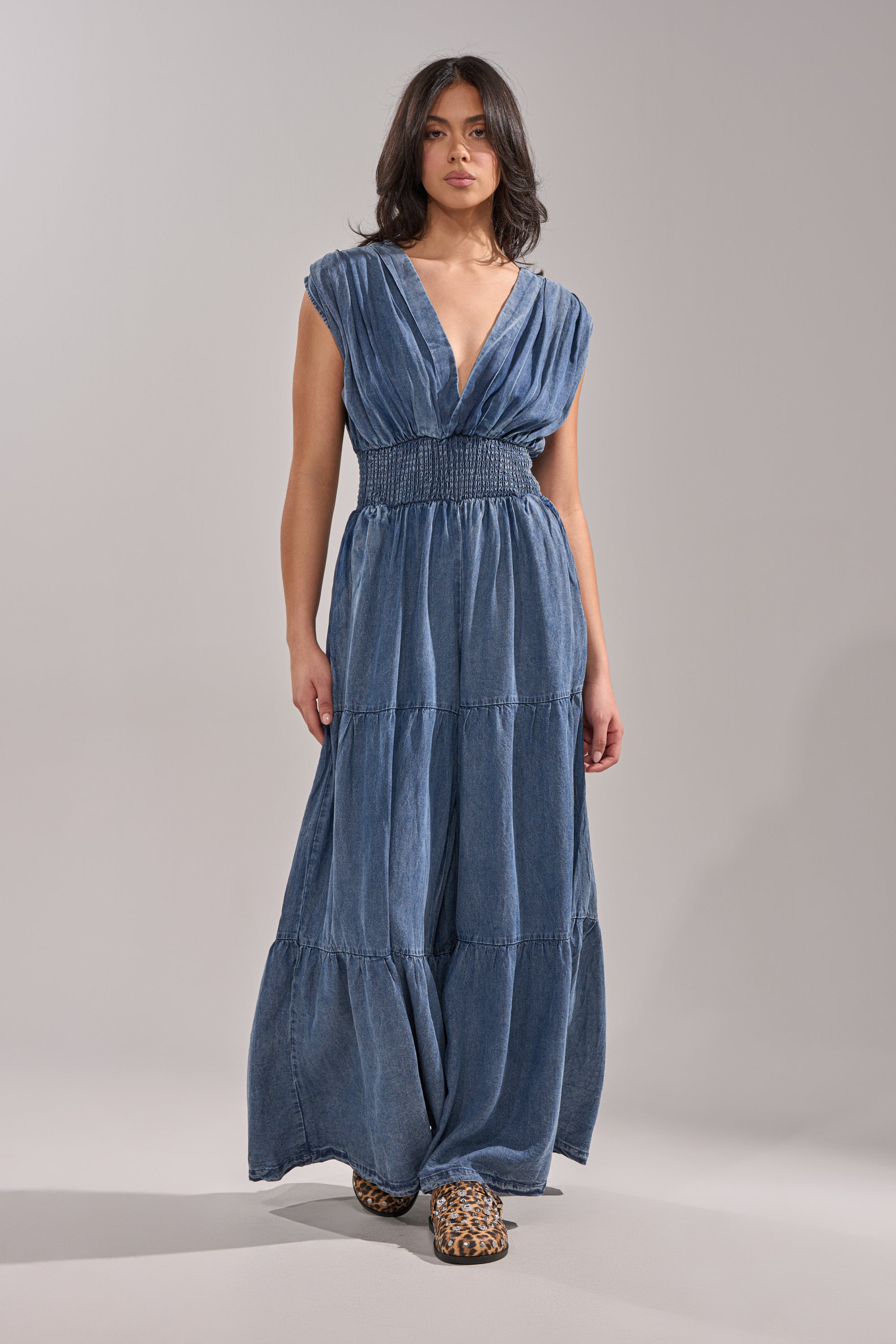 A woman poses against a plain backdrop wearing the KELLY DENIM JUMPSUIT, a sleeveless deep V-neck piece with a shirred waist and tiered skirt, styled casually and paired with leopard print shoes. 3
