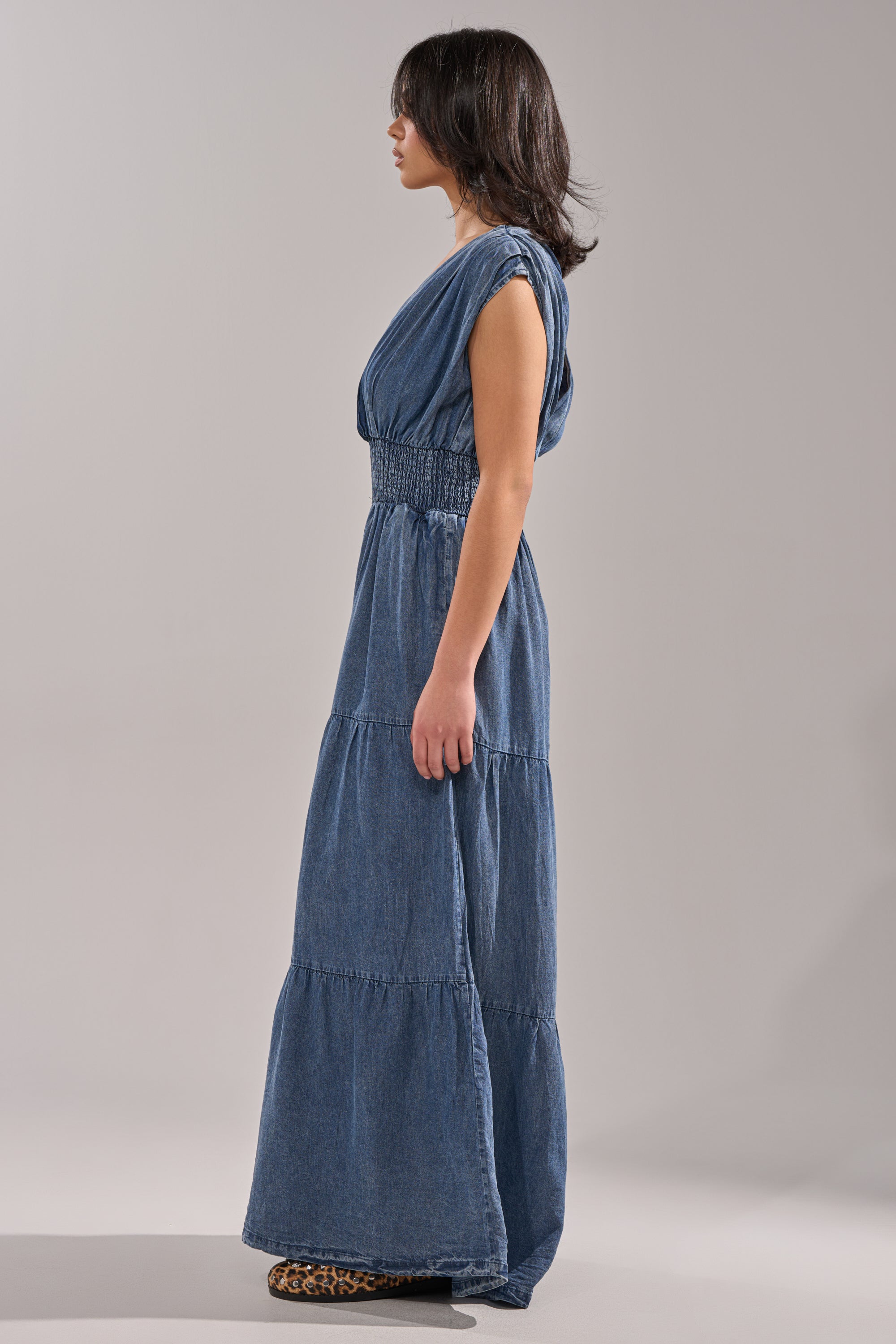 A woman stands in profile against a plain background wearing the KELLY DENIM JUMPSUIT, a sleeveless, ankle-length, tiered blue denim piece with a gathered waist—perfect for casual looks—paired with leopard print shoes. 4