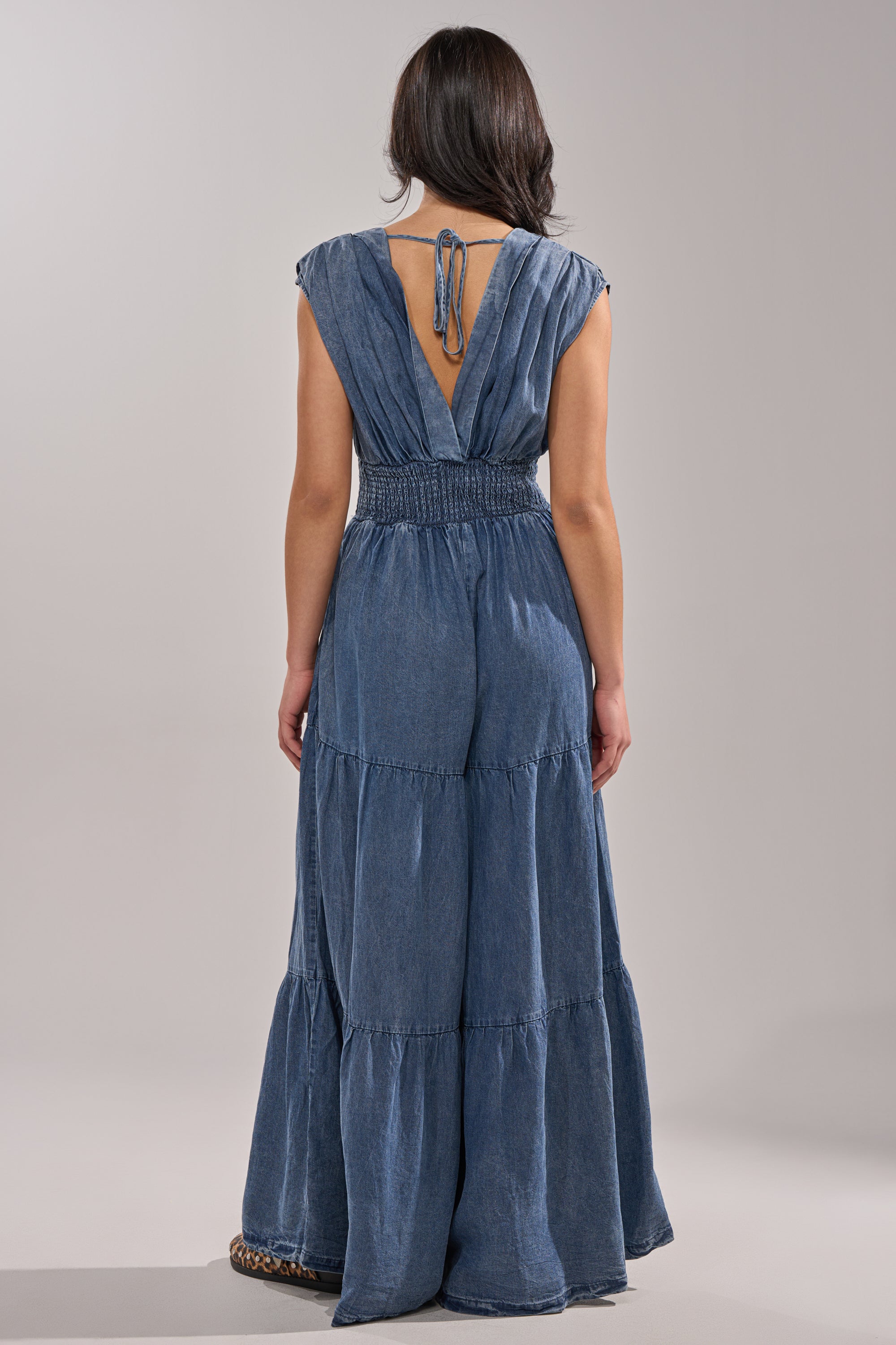 A woman faces away in the KELLY DENIM JUMPSUIT, a sleeveless, floor-length blue piece with a deep V-back, tiered skirt effect, neck tie, and cinched waist. The background is plain and neutral for an effortless casual look. 5