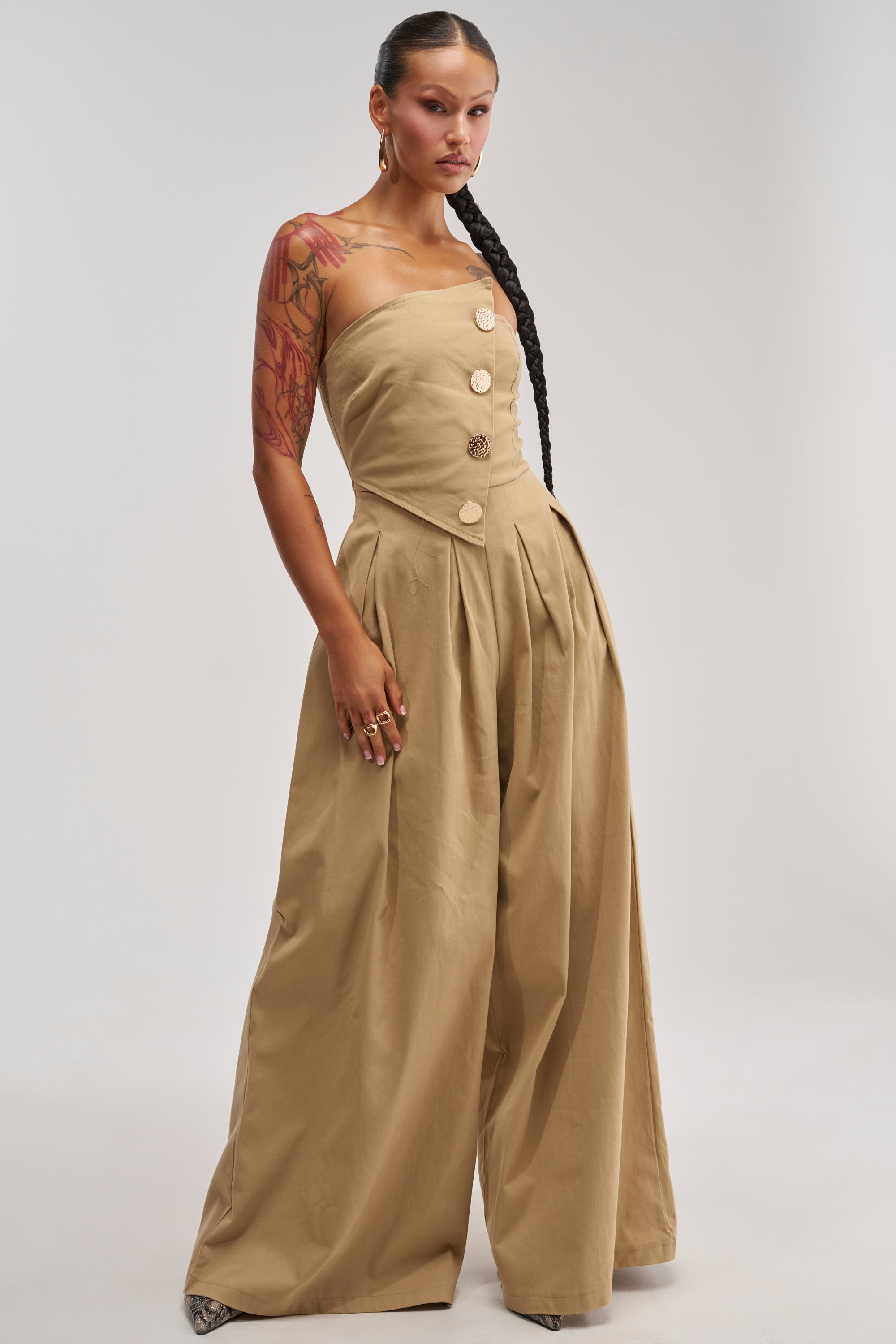 A woman with a long braid and arm tattoos models the MILANO WOVEN JUMPSUIT IN KHAKI, featuring a strapless design with large buttons and wide legs, standing against a plain light background.