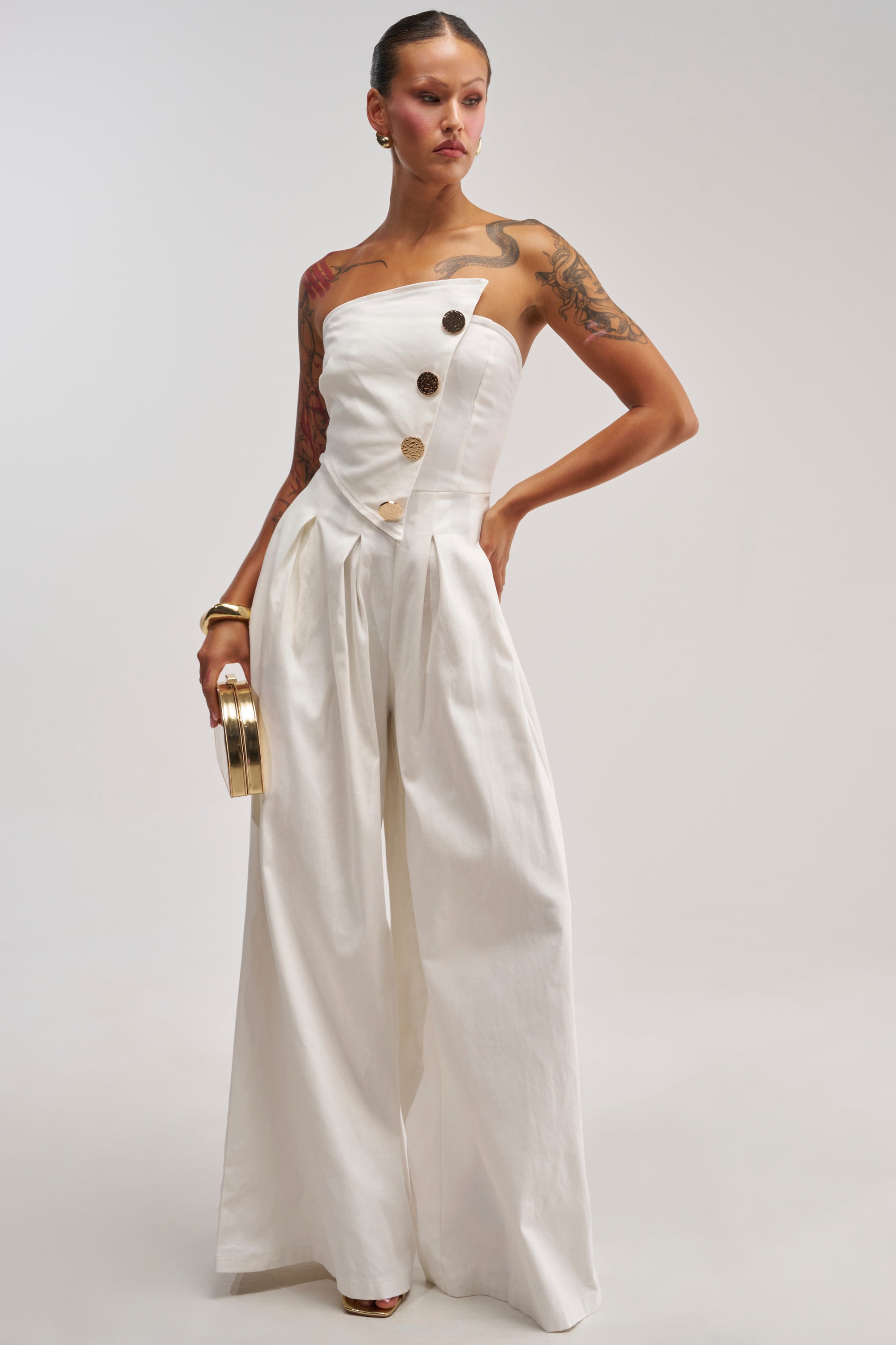 Dressed in the MILANO WOVEN JUMPSUIT IN WHITE—a chic strapless design featuring wide-leg pants and prominent decorative buttons—a woman holds a small gold clutch, accessorized with gold bracelets that highlight her arm tattoos, all against a plain backdrop. This jumpsuit effortlessly fuses contemporary elegance with striking accessories.