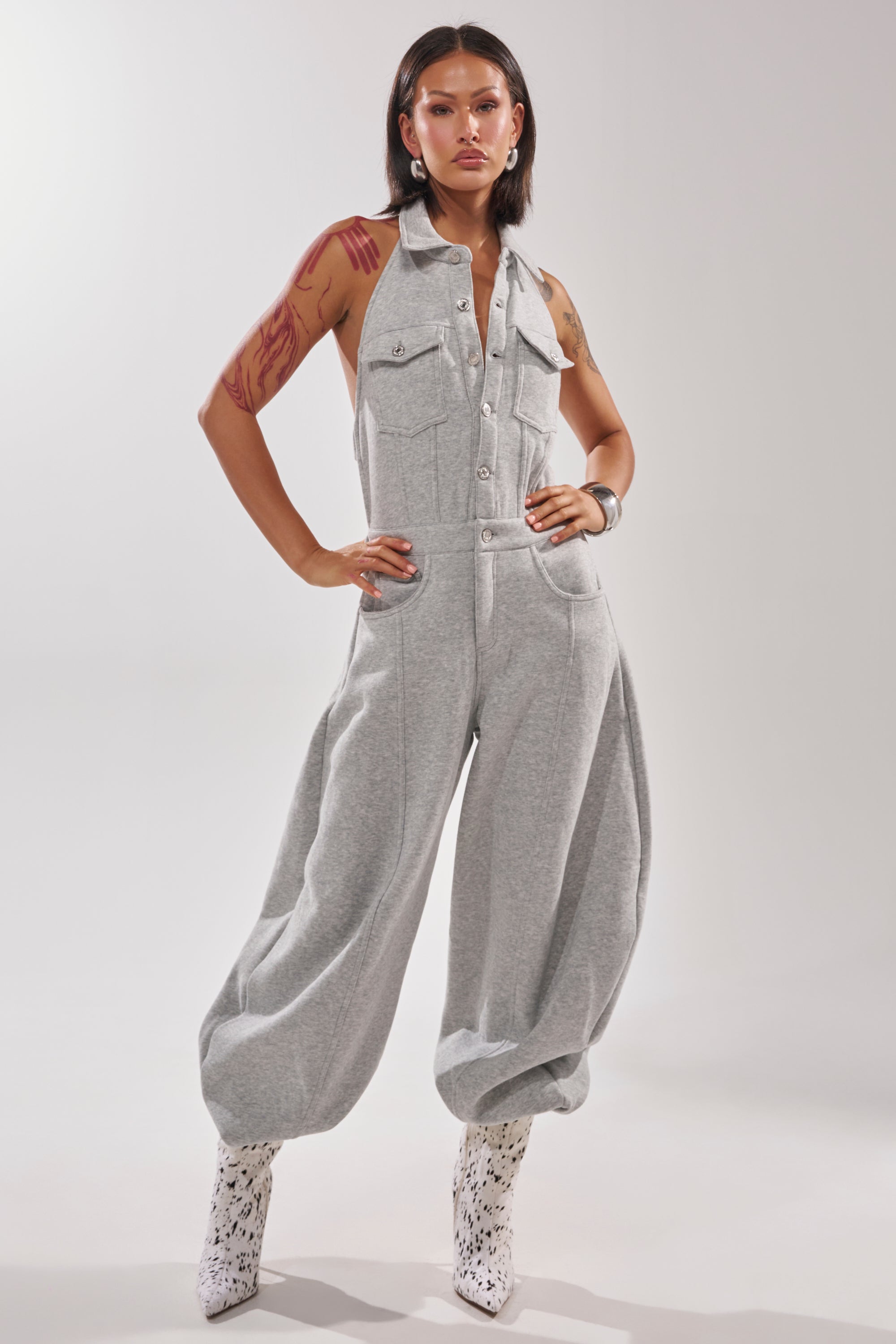 A person with tattoos, dark hair, and hoop earrings poses confidently in the JETSETTER JUMPSUIT IN FRENCH TERRY—a sleeveless gray piece with wide legs and front pockets—paired with white patterned boots against a plain background.