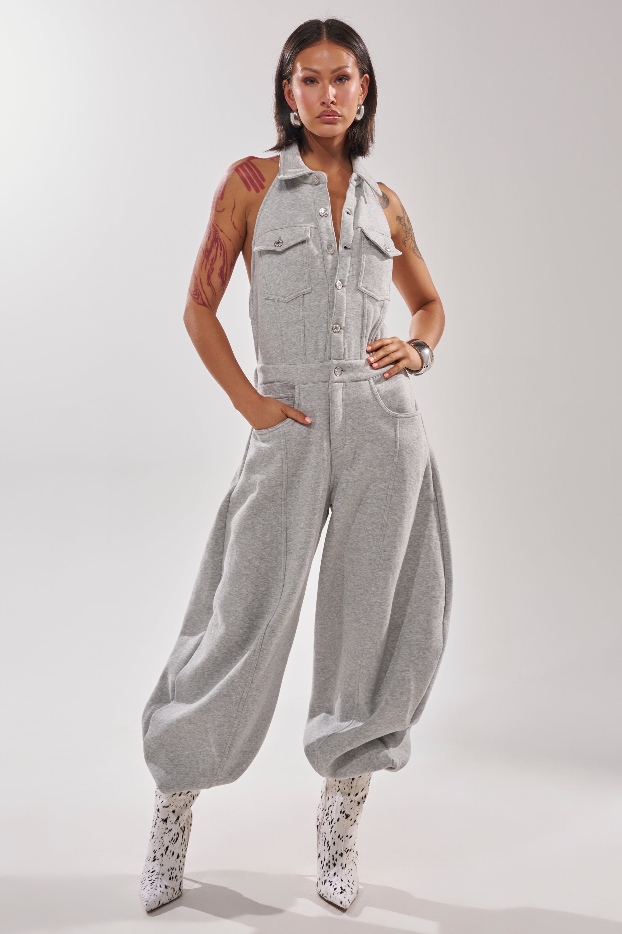 A woman confidently poses in the JETSETTER JUMPSUIT IN FRENCH TERRY, a sleeveless light gray halter jumpsuit with wide legs, paired with white patterned boots and showing her arm tattoos against a plain light background.
