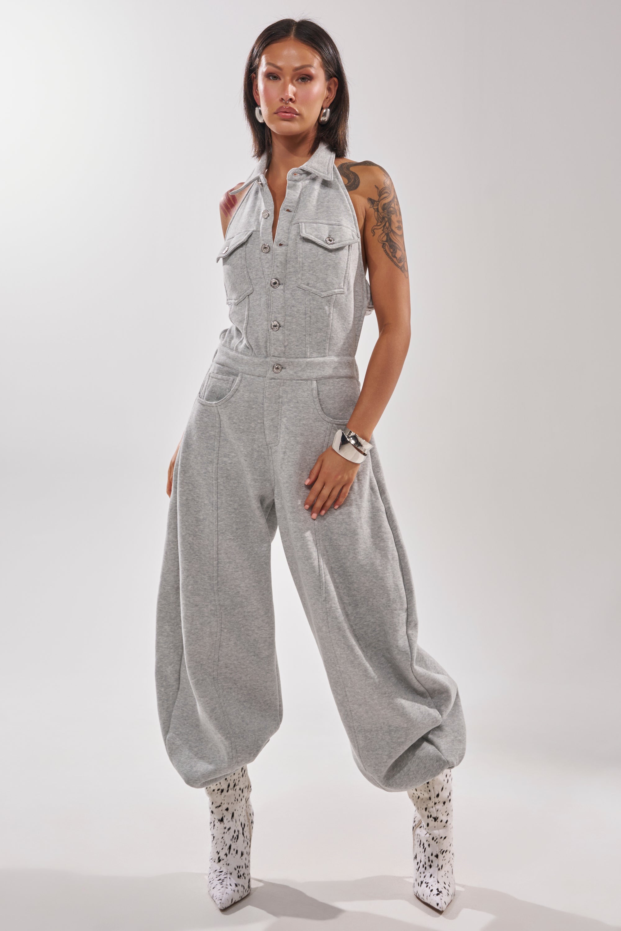 A woman exudes confidence in the JETSETTER JUMPSUIT IN FRENCH TERRY, featuring wide legs and large pockets. She pairs it with white patterned boots, accessorizing with a silver watch and earrings, her long dark hair highlighting visible tattoos.