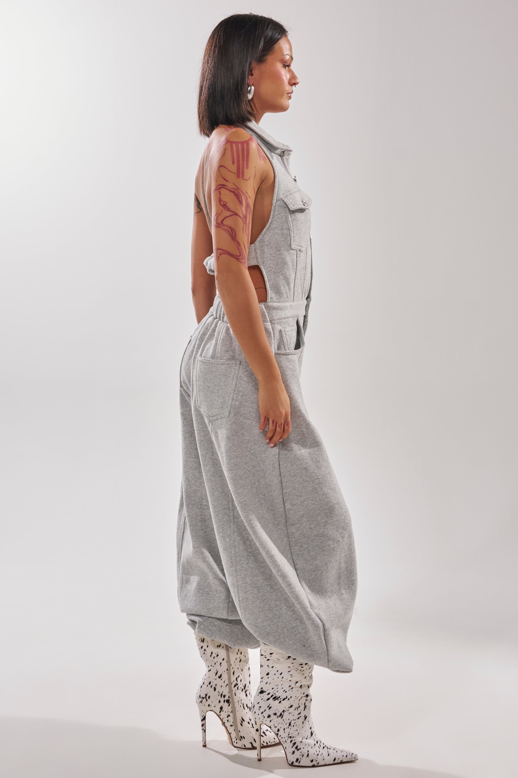 A woman in profile wears the JETSETTER JUMPSUIT IN FRENCH TERRY with white boots featuring a black splatter pattern. She has straight dark hair, a bold red design on her arm, and stands against a plain light background.