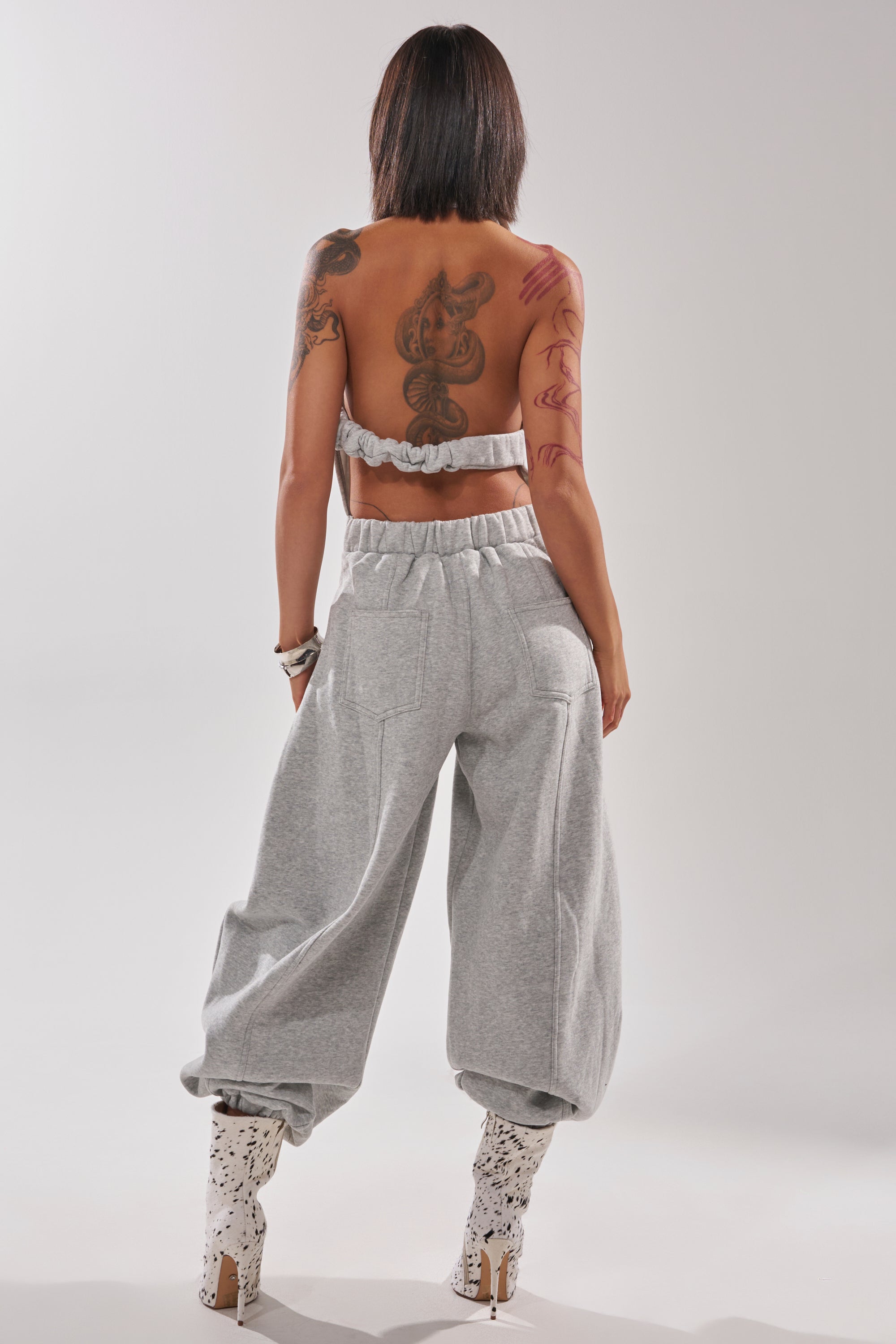 A woman with shoulder-length straight dark hair faces away, revealing back tattoos. She wears the strapless JETSETTER JUMPSUIT IN FRENCH TERRY, paired with white heeled boots featuring black patterns. The background is plain and light.