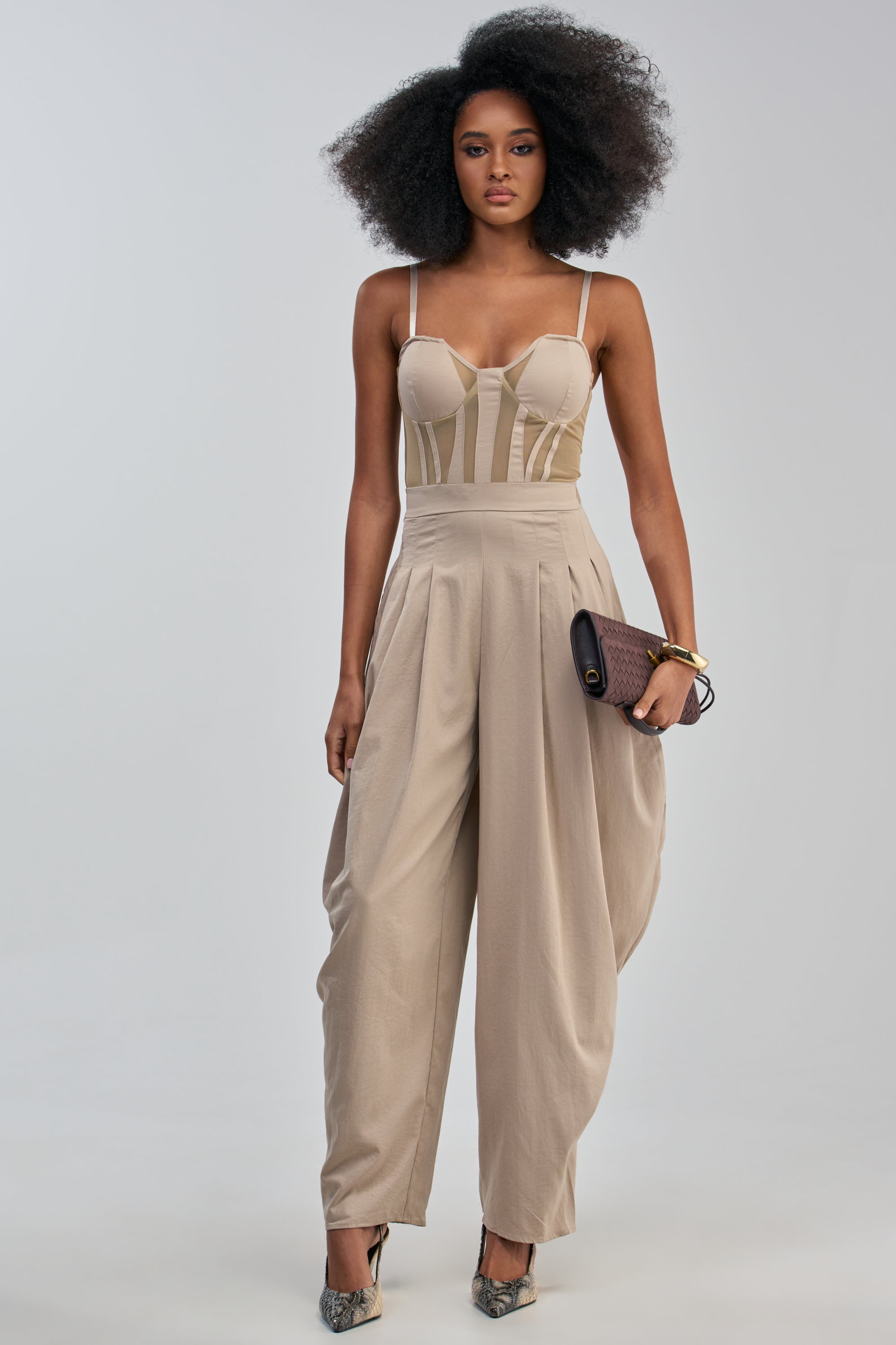 A woman with voluminous curly hair poses confidently against a plain background wearing the NO FUNNY BUSINESS CORSET JUMPSUIT, which features a flattering sweetheart neckline, dramatic balloon pant legs, and a chic khaki hue. She accessorizes with a dark clutch and pointed heels.