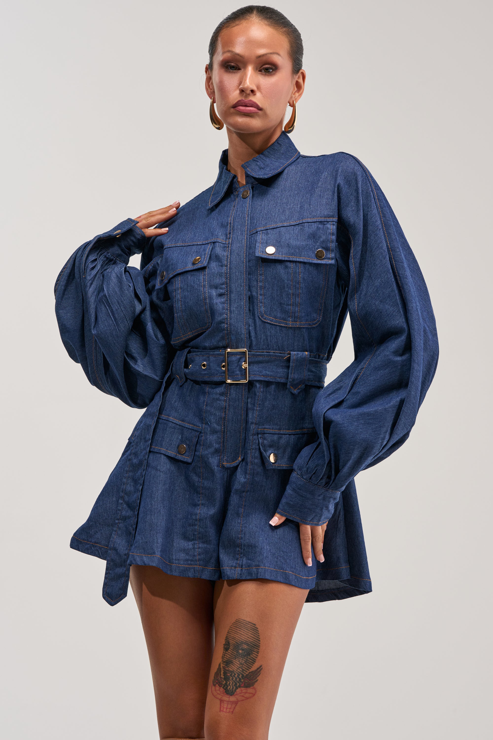 A woman wears the CITYSCAPE BELTED DENIM ROMPER, featuring dark denim fabric, long puffed sleeves, a belt at the waist, and front pockets. She accessorizes with hoop earrings and displays a skull tattoo on her thigh.