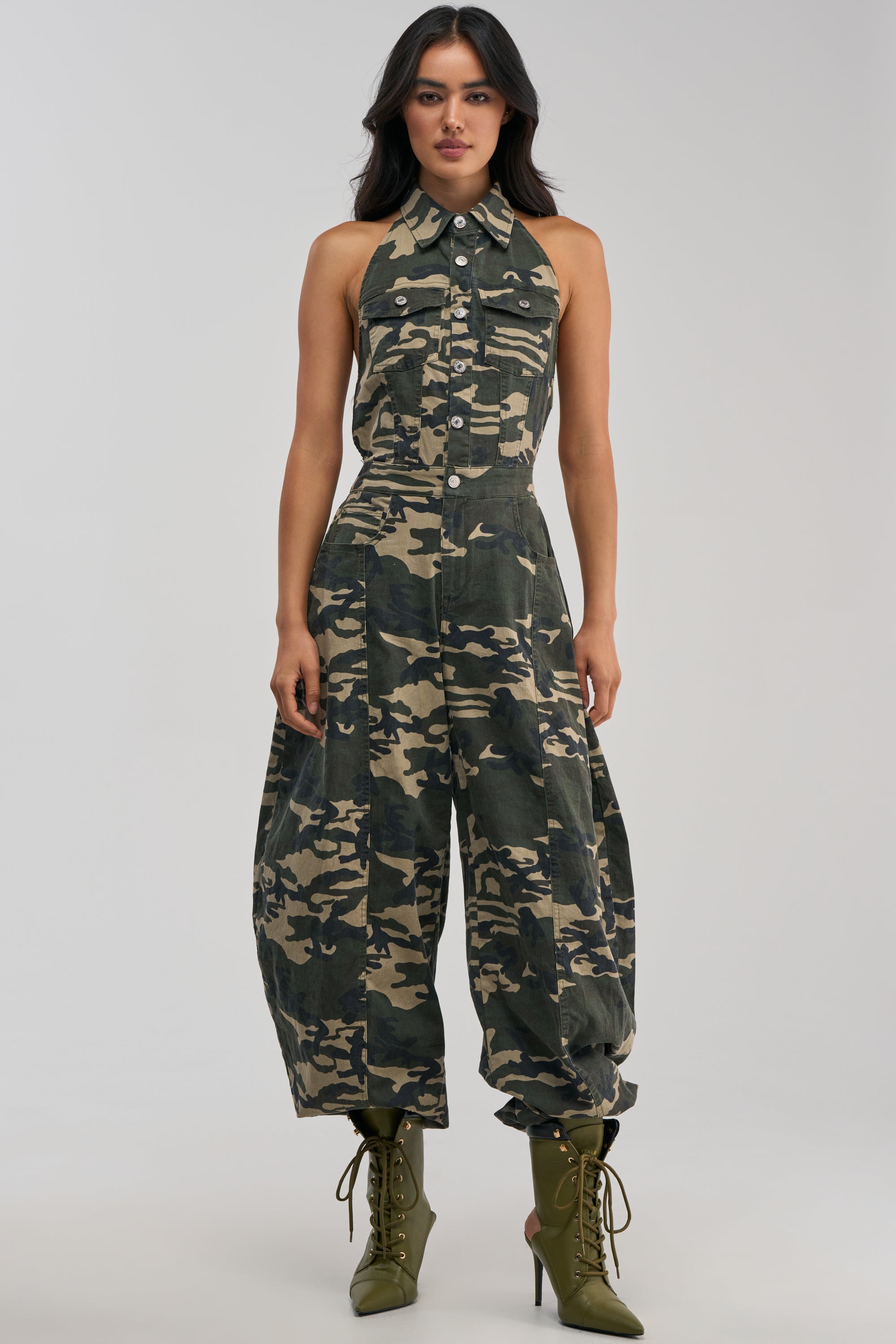 Model stands against a plain light background wearing the COUNT ME IN CAMO HALTER JUMPSUIT—a sleeveless, button-up camo halter design with wide legs—her hands relaxed by her sides and paired with olive green lace-up boots, showcasing a bold streetwear look that’s perfect for festival season.