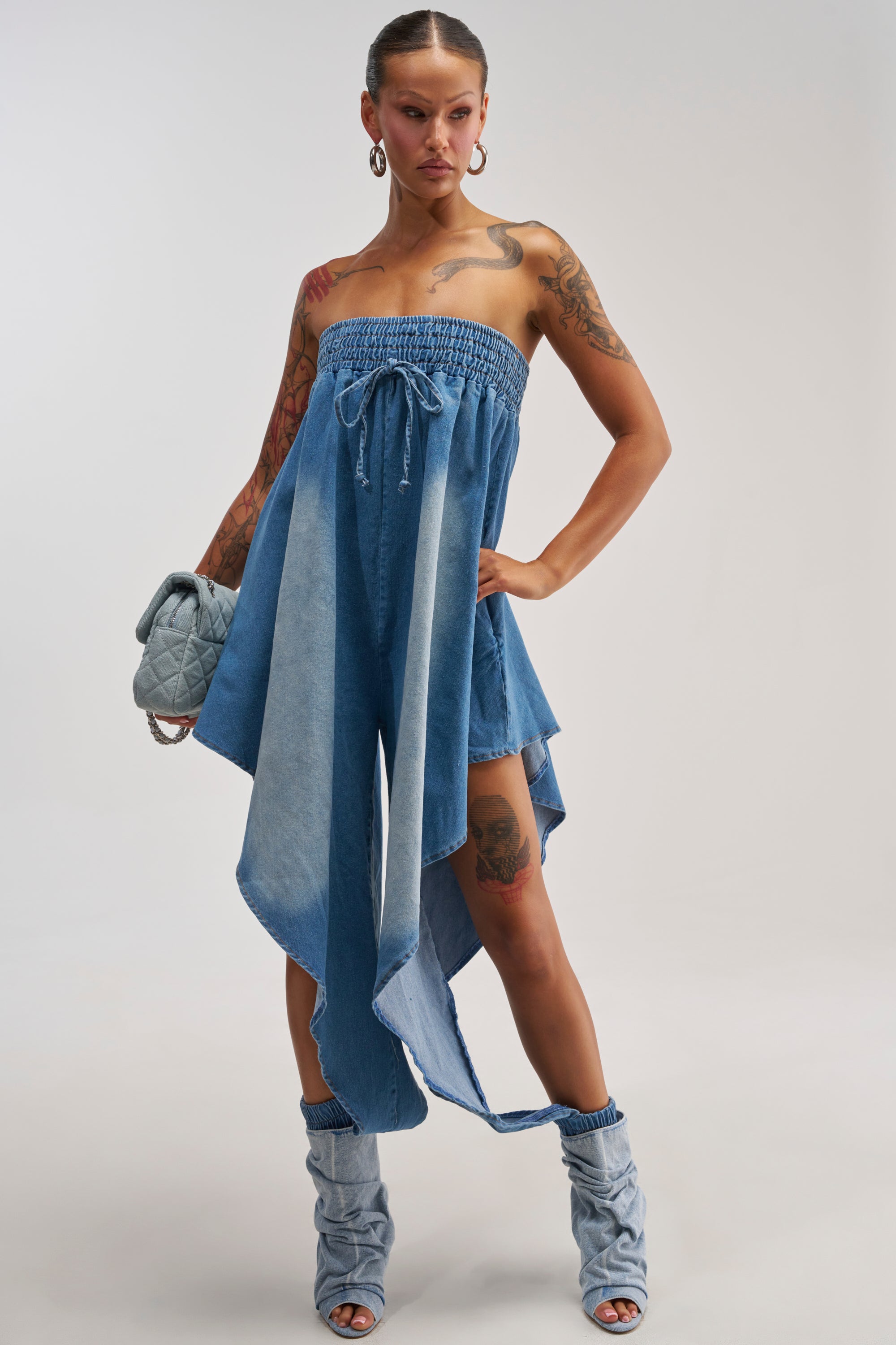 A person with tattoos poses confidently against a plain white background wearing the DIFFERENT FROM THE REST DENIM JUMPSUIT—strapless with an asymmetrical cut—paired with matching denim boots, hoop earrings, and a quilted purse.