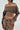 A person in a BROKE MY HEART OFF THE SHOULDER SWEATSHIRT, styled in a cropped silhouette and paired with a brown denim high-waisted skirt, accented by hoop earrings, stands confidently with one hand on their hip against a plain background.