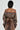 Facing away, a person with short dark hair is wearing the BROKE MY HEART OFF THE SHOULDER SWEATSHIRT in brown, which features a lace-up back closure. They have paired it with a brown corset belt and high-waisted, textured brown pants. The scene is set against a plain white background.