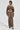 A person stands against a plain background, wearing the BROKE MY HEART OFF THE SHOULDER SWEATSHIRT in brown featuring a lace-up back closure, styled with a matching long skirt that includes a high corset-like waistband and paired with chunky black and white sandals.