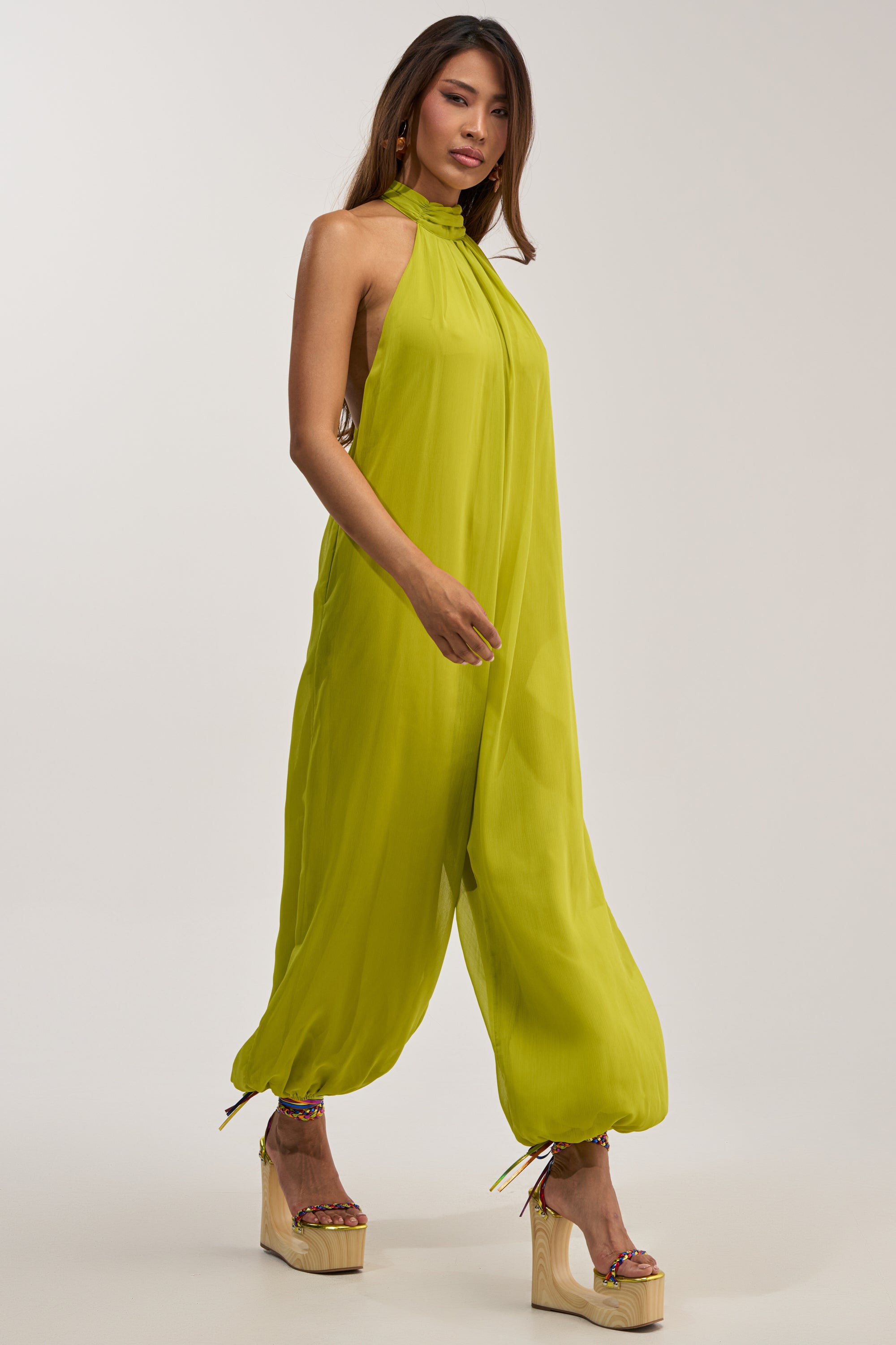 A woman models the PLUS ONE CHIFFON JUMPSUIT IN CHARTRUESE, a vibrant yellow-green halter-neck jumpsuit featuring loose, gathered ankles and paired with platform wedge sandals. Perfect for summer evenings, this chic chiffon design delivers a blend of bold style and effortless comfort against a simple light backdrop.