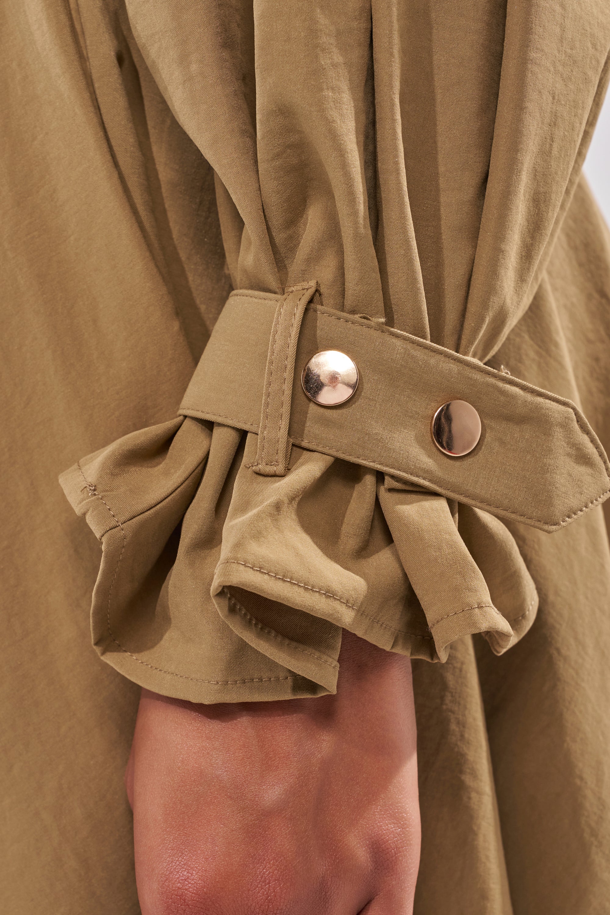 A close-up of a person’s arm wearing the CANT STOP WONT STOP BUTTON DOWN, a tan cropped long-sleeve shirt with a decorative cuff strap, two shiny gold buttons, and ruffled details at the sleeve edge. 9