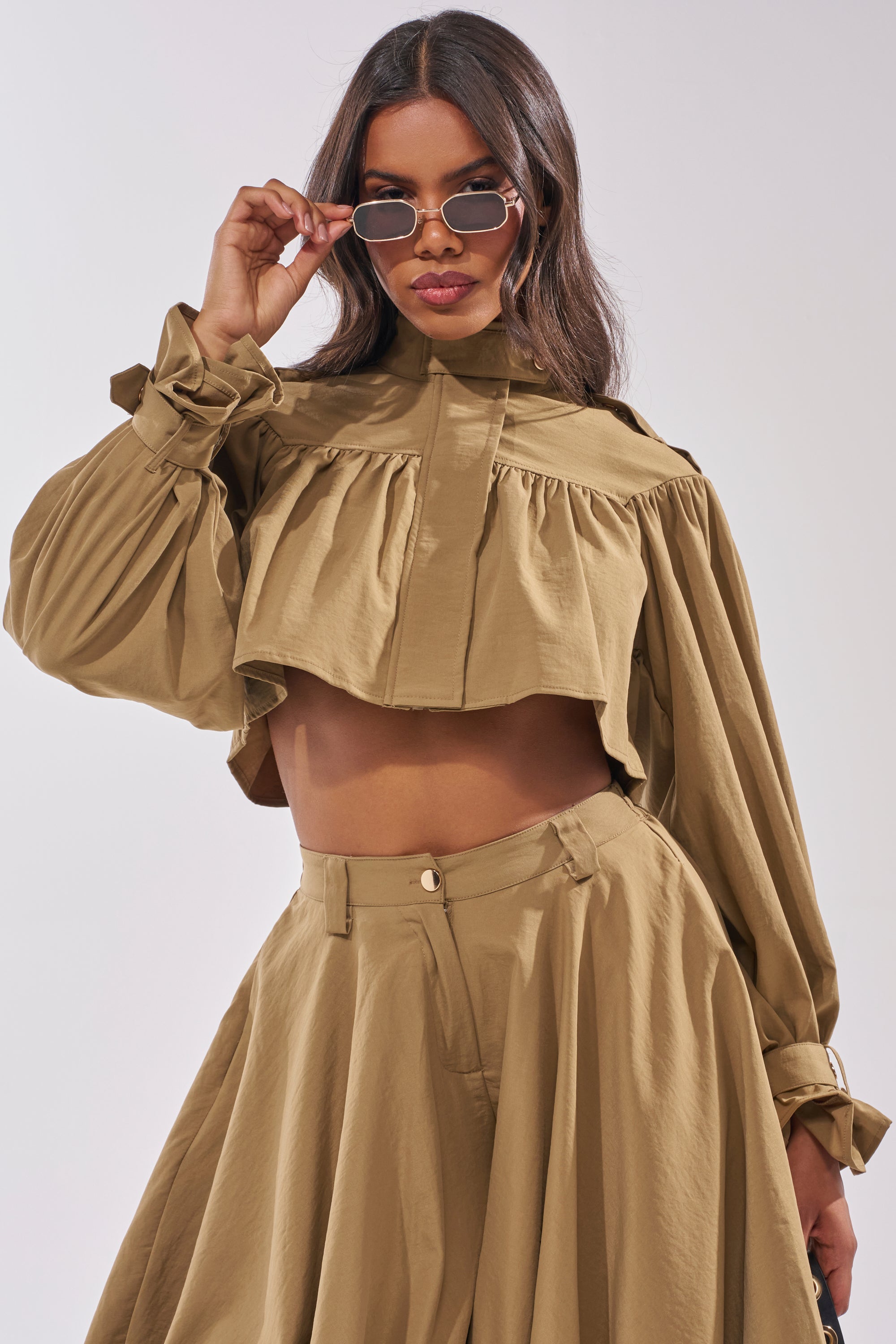 A woman poses confidently against a plain background, wearing the CANT STOP WONT STOP BUTTON DOWN top styled with a matching high-waisted skirt and sunglasses, lifting her shades with one hand. 2