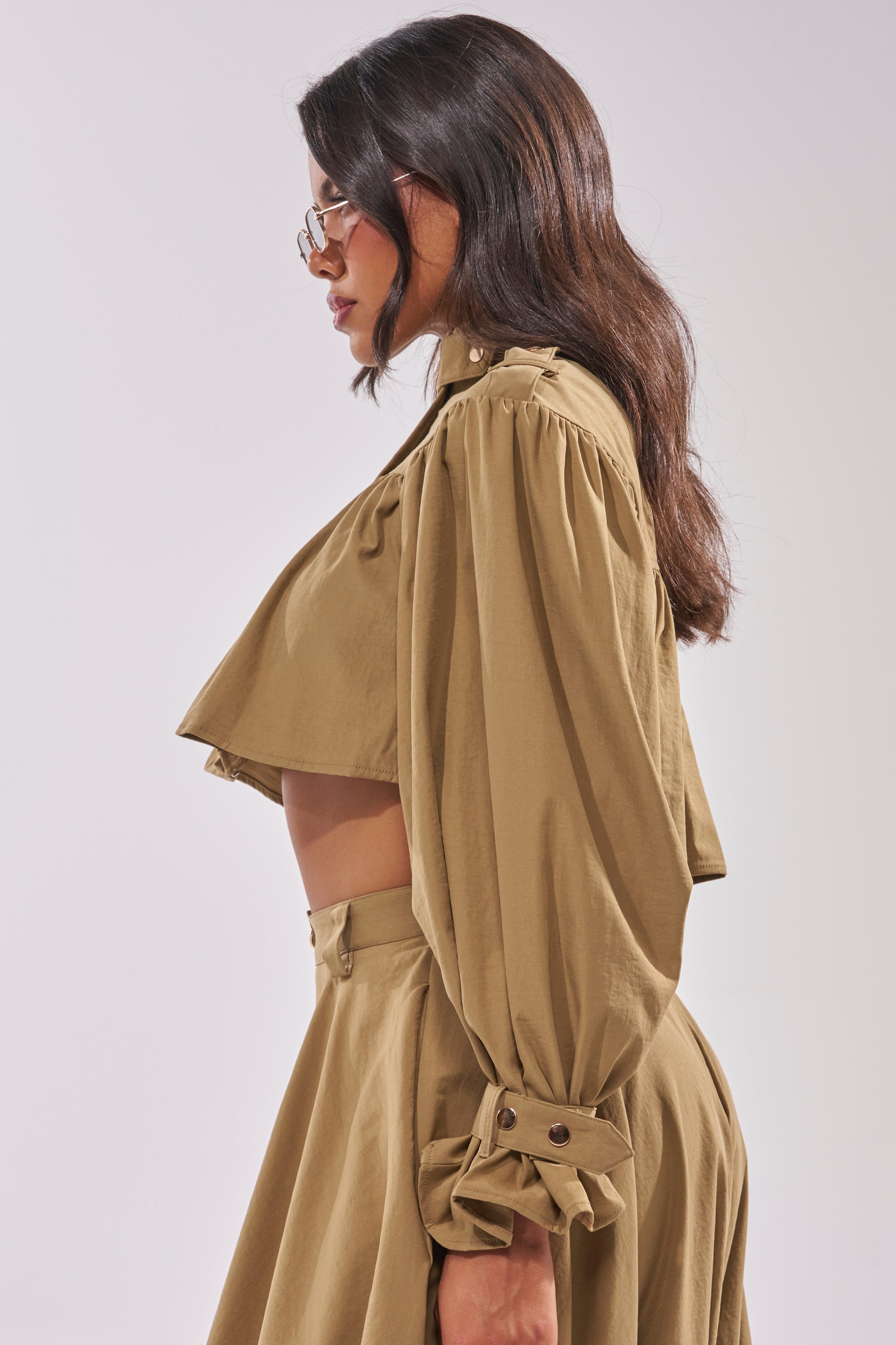 A woman in profile with wavy brown hair and glasses wears the CANT STOP WONT STOP BUTTON DOWN—a light brown, long-sleeve, pleated top with puffy sleeves—paired with a matching high-waisted skirt against a plain light background. 3
