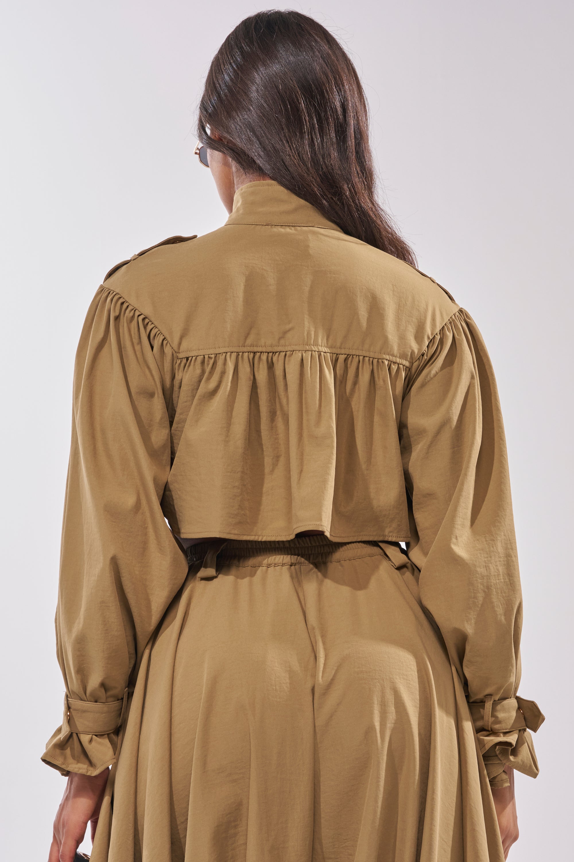 A person with long brown hair is seen from the back wearing the CANT STOP WONT STOP BUTTON DOWN—a light brown, long-sleeved dress with a cropped silhouette, gathered fabric, and jacket details. 4