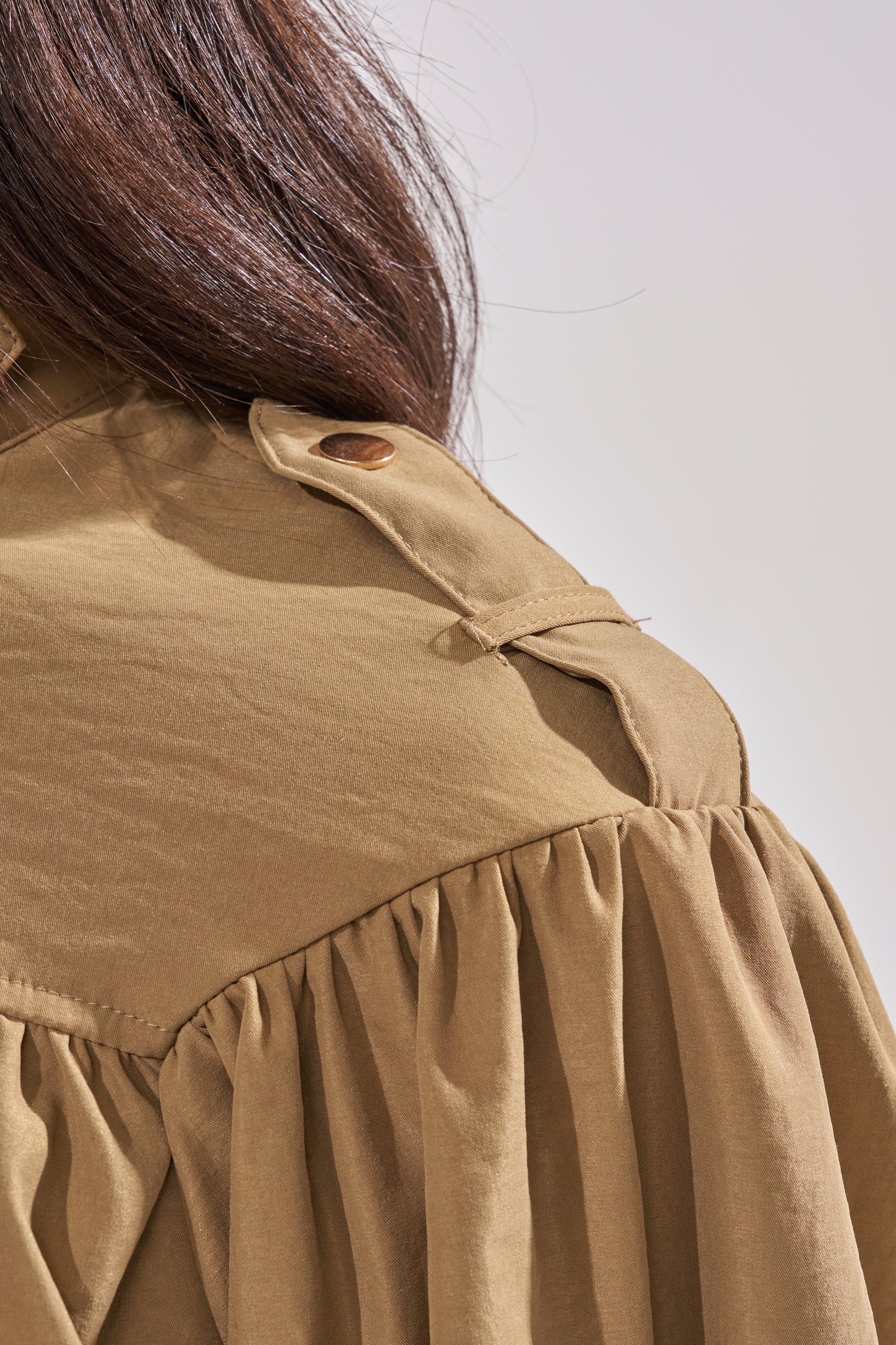 Close-up of a person wearing the CANT STOP WONT STOP BUTTON DOWN—a light brown, utilitarian-chic top with gathered fabric, an epaulet on the shoulder, and a cropped silhouette. The person's long brown hair is partially visible. 8