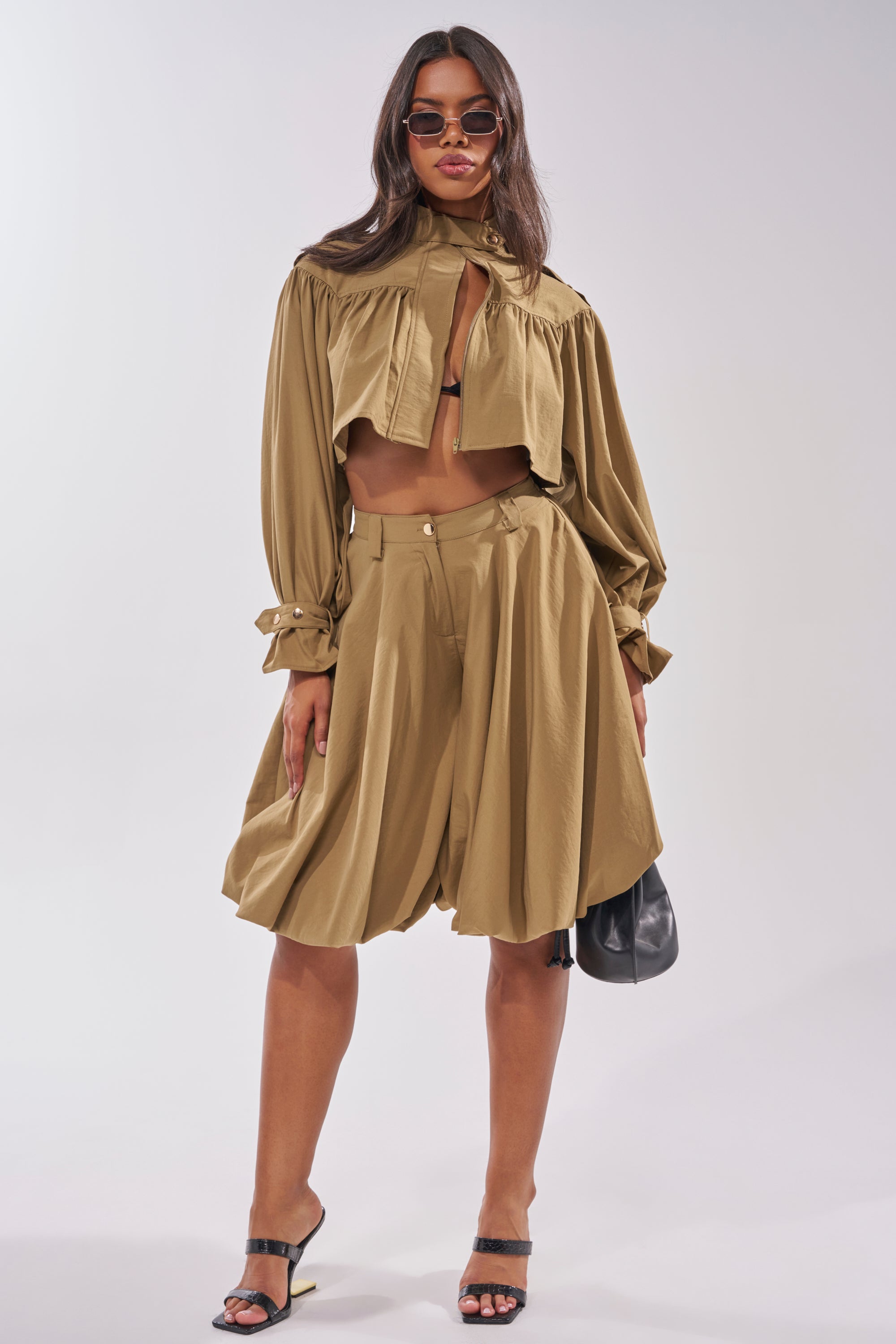 A woman stands confidently in a loose-fitting tan cropped jacket, CANT STOP WONT STOP BERMUDA SHORT with a high-rise waistband, black bralette, heeled sandals, sunglasses, and holding a small black bag against a light background. 0
