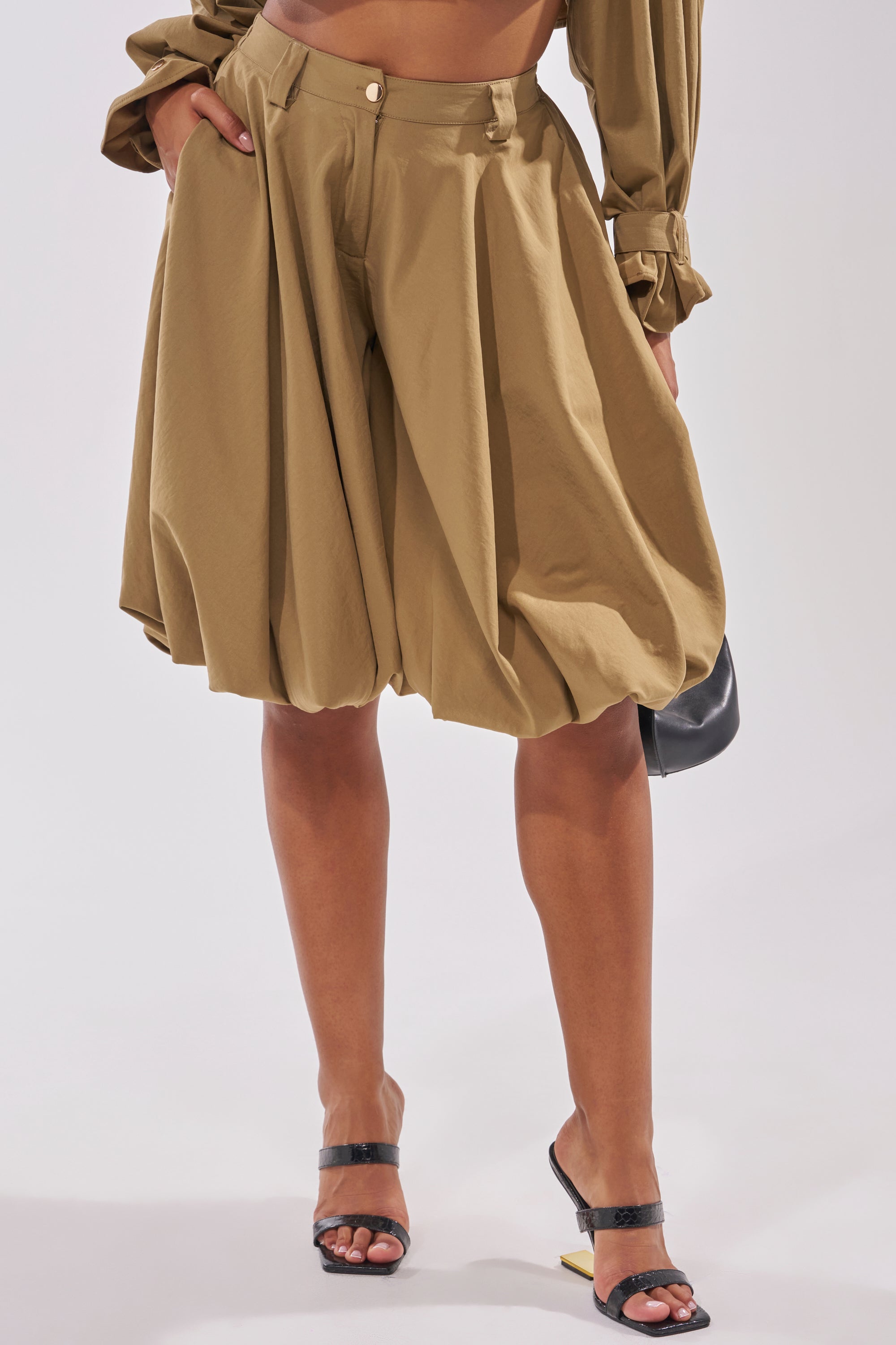 A person is seen from the waist down wearing the CANT STOP WONT STOP BERMUDA SHORT in tan with a matching long-sleeve top, black heeled sandals, and holding a small black bag. 2
