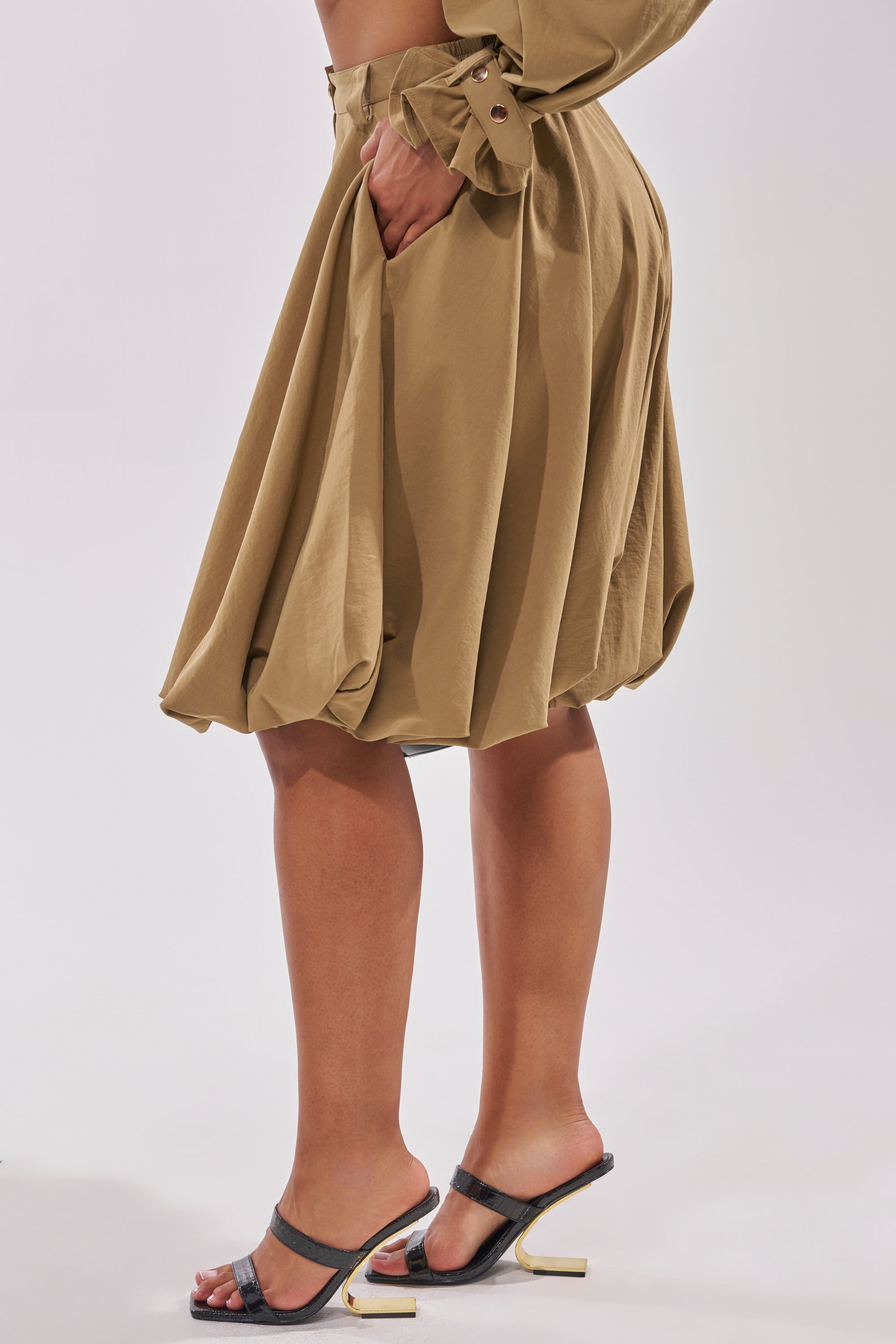 A person models the CANT STOP WONT STOP BERMUDA SHORT in tan, styled with black heeled sandals and a hand in their pocket, standing against a plain light background. 4