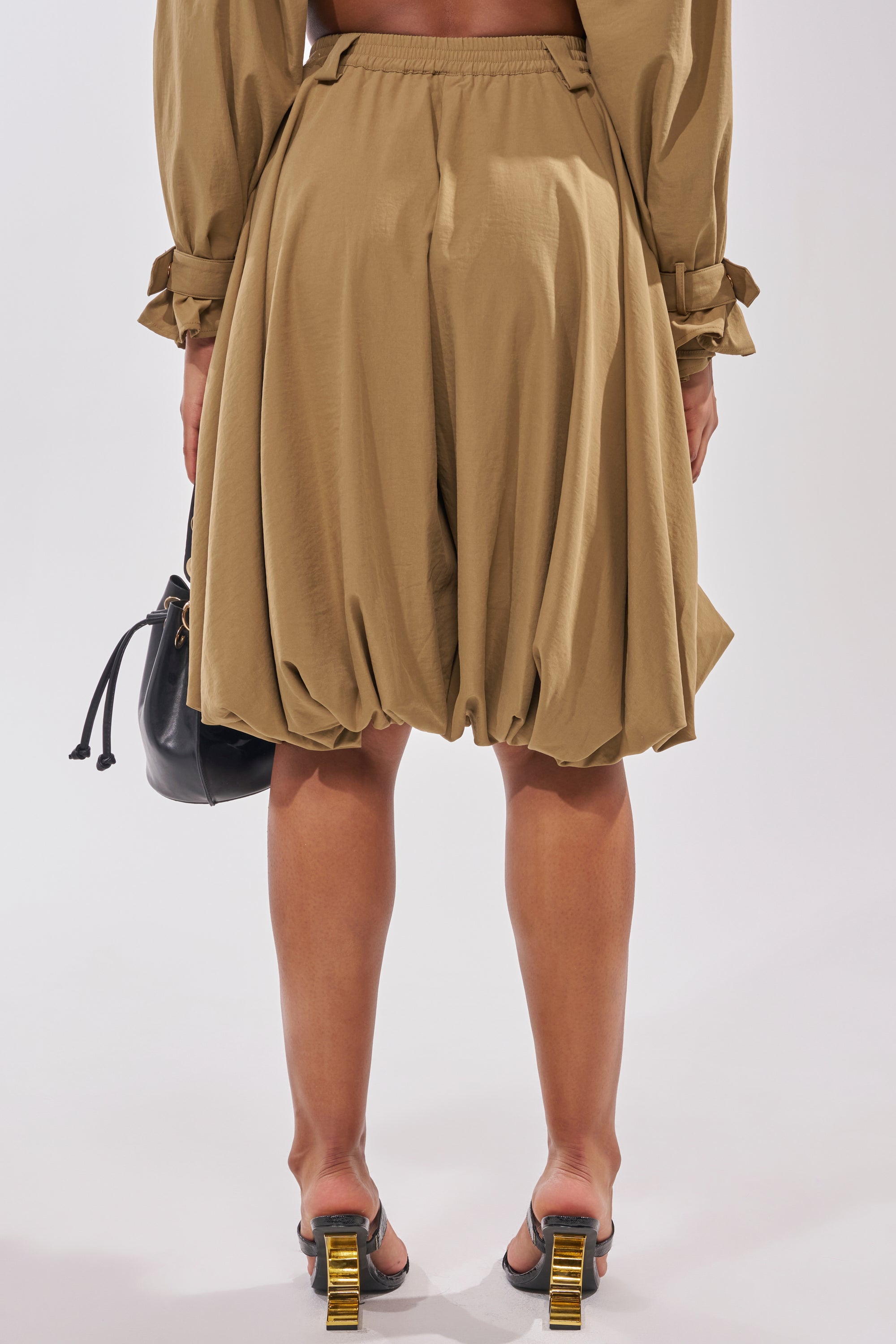 A person faces away, wearing the CANT STOP WONT STOP BERMUDA SHORT in tan with a matching long-sleeve top, heeled sandals with gold accents, and holding a black drawstring bag against a plain light background. 5
