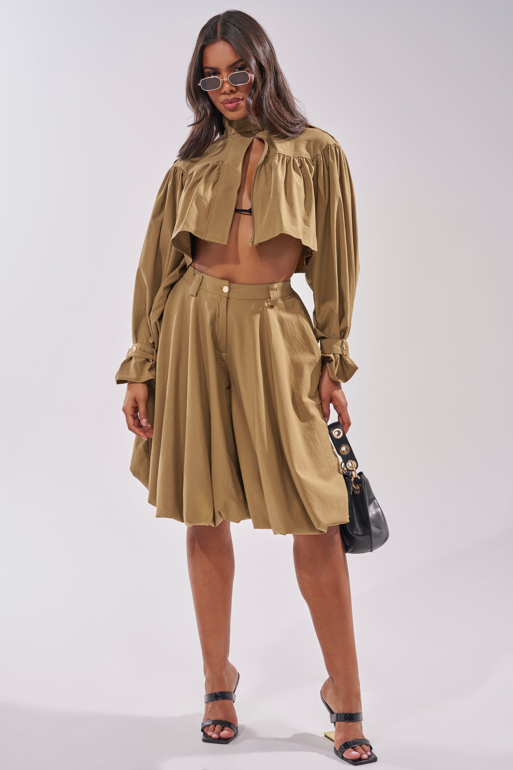 A woman poses in a tan, long-sleeve cropped blouse and matching CANT STOP WONT STOP BERMUDA SHORT with a high-rise waistband, paired with black sandals, sunglasses, and a black handbag against a plain background. 6