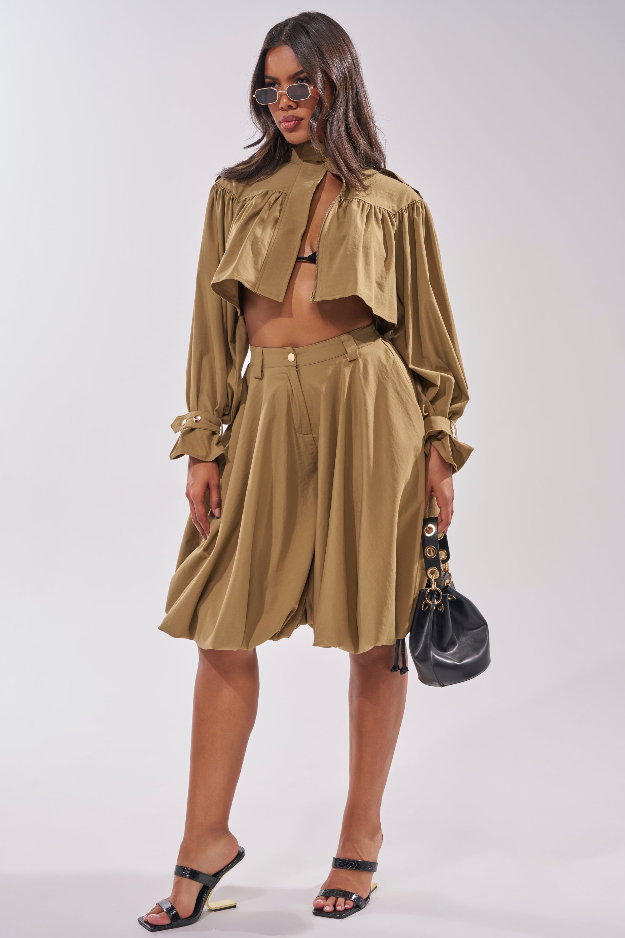 A woman wearing CANT STOP WONT STOP BERMUDA SHORTS in olive green, a matching cropped jacket, black bra, black heeled sandals, sunglasses, and holding a small black handbag poses against a plain background. 7