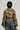 Seen from the back, a person in the AKIRA Label HIGH EXPECTATIONS RUCHED BOLERO, featuring a green camo print and ruched sleeves, is paired with blue jeans adorned with patches on the back pockets. Their short black hair and small bag in hand complement their stance against a plain gray backdrop.