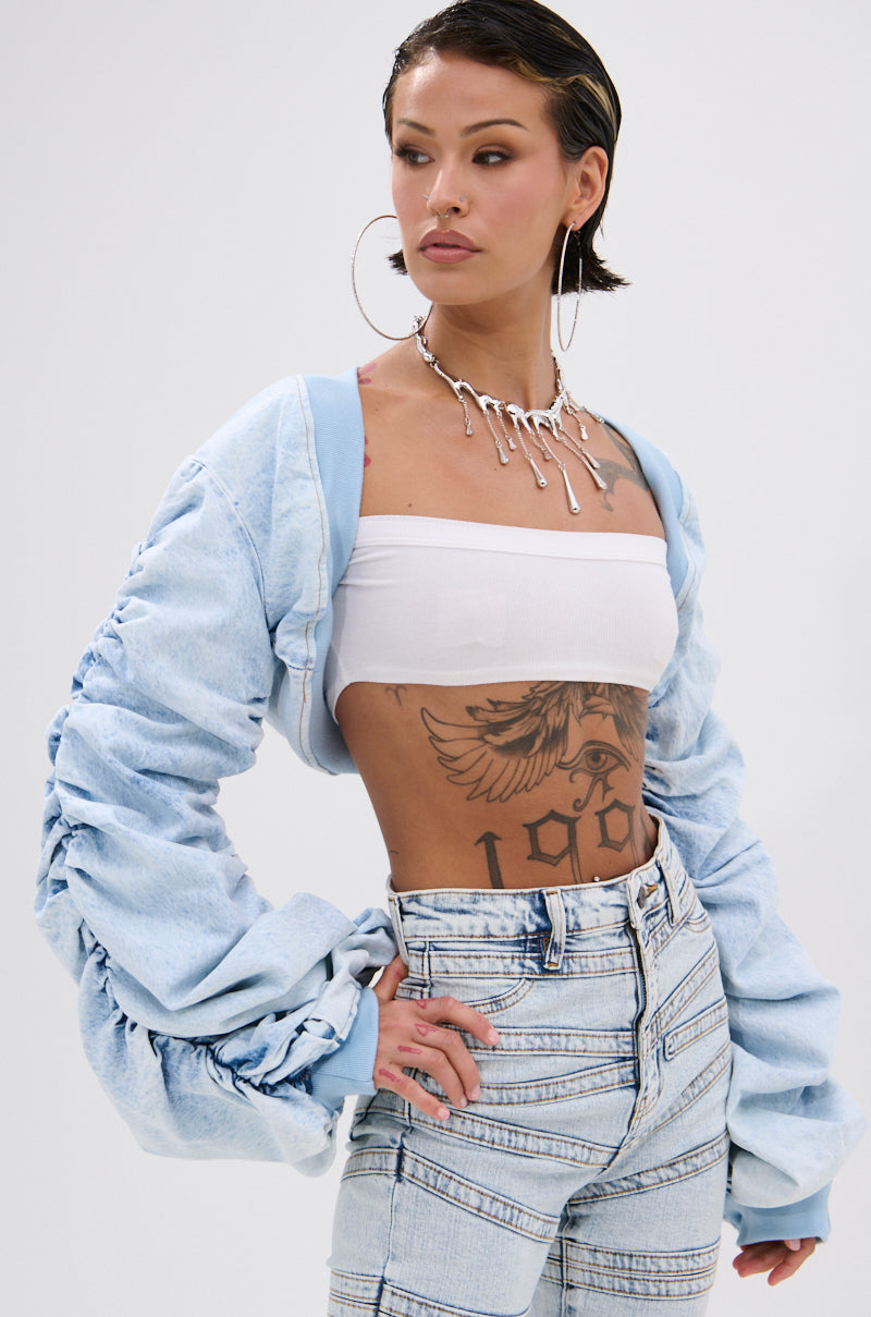 A tattooed woman wears a white bandeau top, plaid high-waisted jeans, large hoop earrings, a chunky necklace, and the DANI RUCHED DENIM BOLERO with ruched sleeves, posing confidently against a plain background.