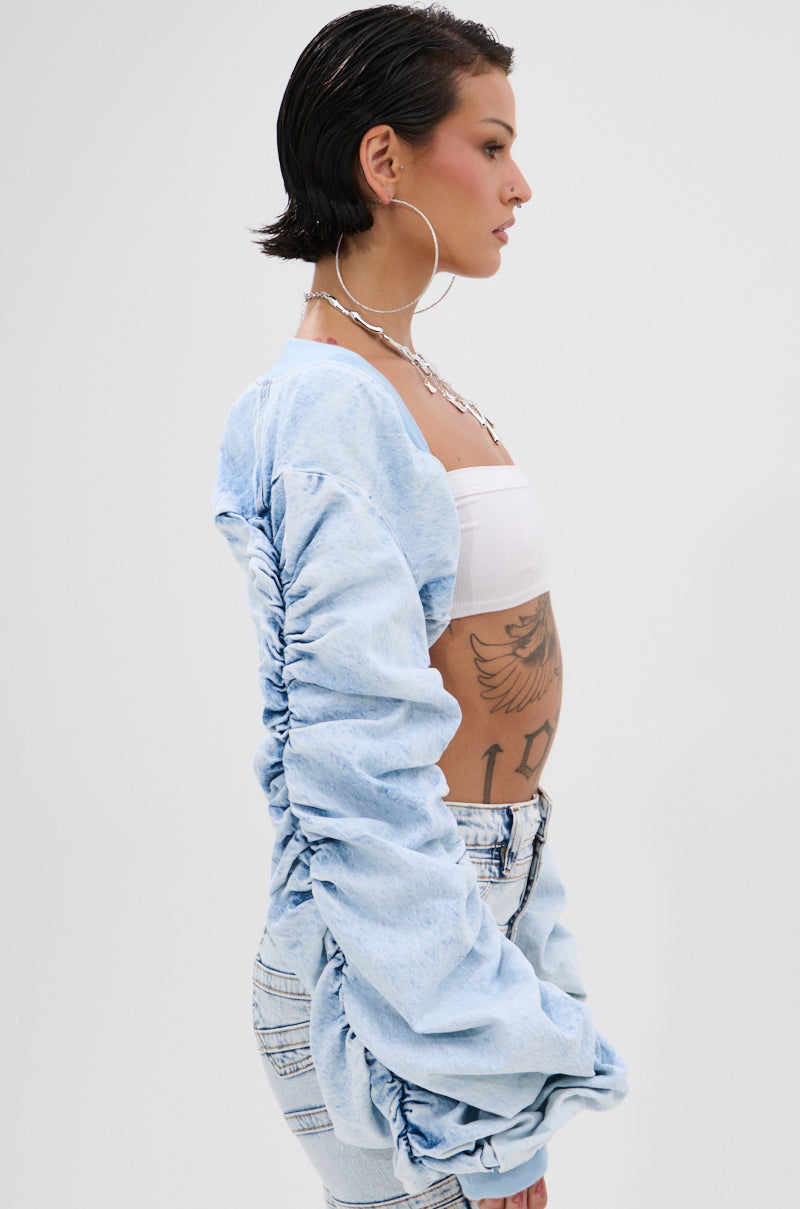 A person with short dark hair and large hoop earrings wears the DANI RUCHED DENIM BOLERO over a white bandeau top and light wash jeans, showing tattoos on their side and arm against a plain white background.