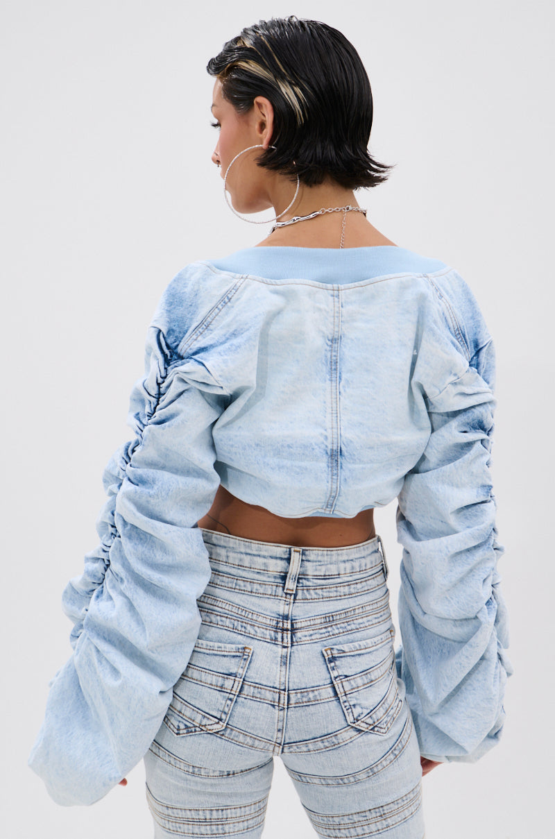 Seen from behind, a person with short, dark, wet-look hair wears the DANI RUCHED DENIM BOLERO—a light denim crop top with ruched long sleeves—paired with matching high-waisted jeans against a plain, light background for an easy streetwear look.