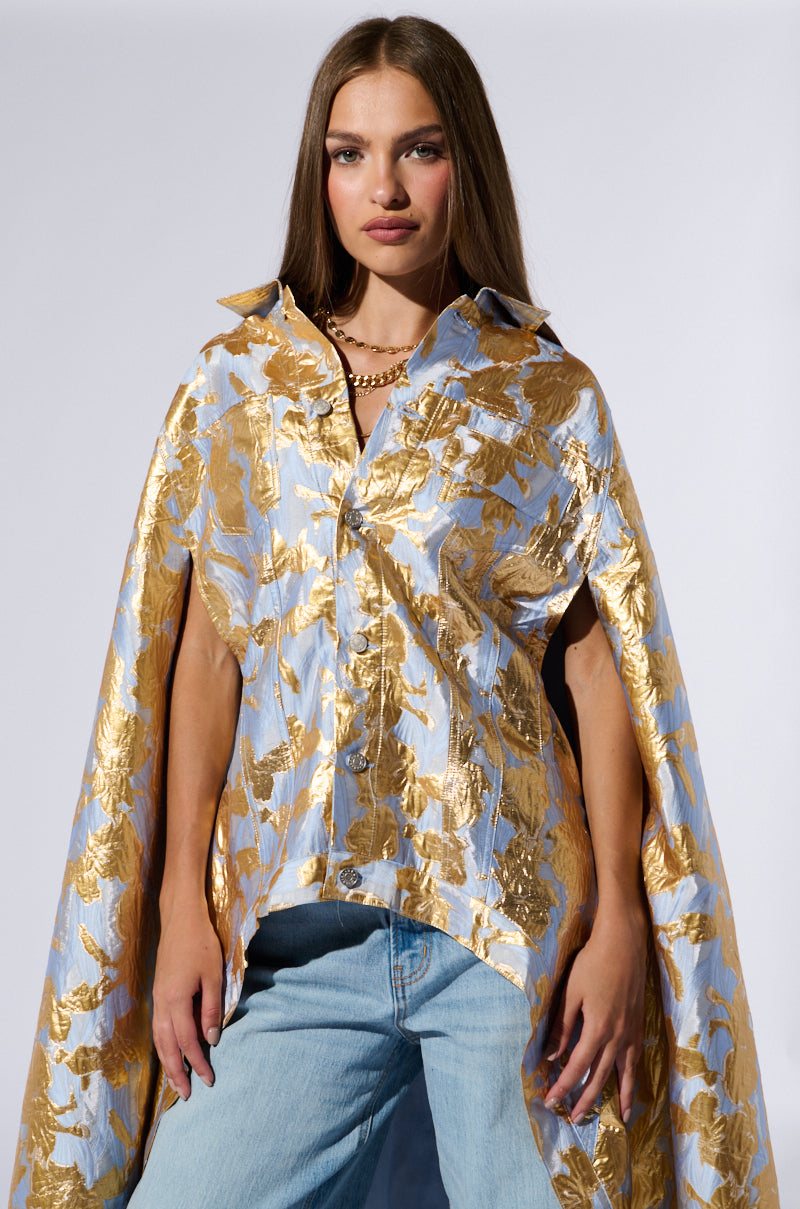 A woman with long hair is standing against a plain background, wearing the OASIS HIGH LOW BROCADE CAPE over light blue jeans. The high-low cape features a striking blue and gold pattern that beautifully complements her confident expression. She has accessorized with a gold necklace. 0
