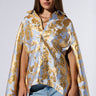 A woman with long hair is standing against a plain background, wearing the OASIS HIGH LOW BROCADE CAPE over light blue jeans. The high-low cape features a striking blue and gold pattern that beautifully complements her confident expression. She has accessorized with a gold necklace.