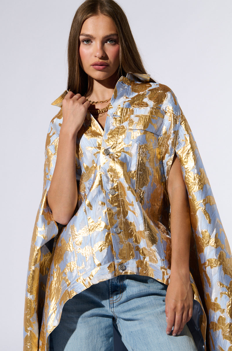 A woman with long brown hair poses confidently, wearing an OASIS HIGH LOW BROCADE CAPE in a metallic gold and light blue pattern. She pairs the high-low silhouette cape with light blue jeans and stands against a neutral background. 2