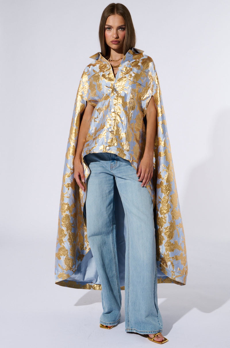 A woman stands against a plain background wearing an OASIS HIGH LOW BROCADE CAPE, which features a shiny, gold-and-purple metallic brocade pattern. She pairs it with light blue, wide-legged jeans. Her straight, shoulder-length brown hair frames her face as she looks directly at the camera in a relaxed pose with her hands by her sides. 3