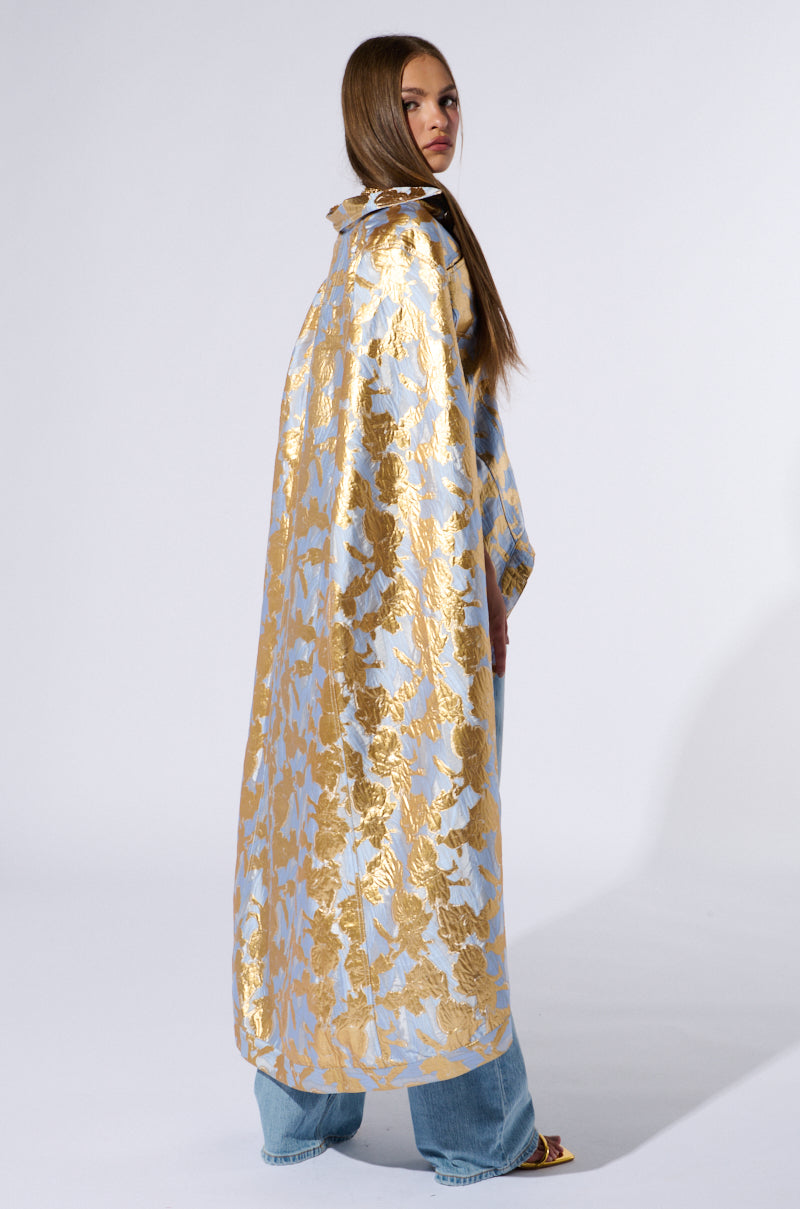 A person with long hair stands sideways, showcasing an OASIS HIGH LOW BROCADE CAPE adorned with a metallic gold and silver pattern. They complement the cape with blue jeans and open-toed shoes, posing against a plain white background. 5