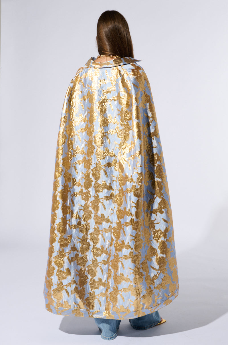 A person with long brown hair stands facing away, showcasing the OASIS HIGH LOW BROCADE CAPE, which features a stunning blue and gold floral metallic brocade pattern. The plain white background highlights the intricate design and the shimmering fabric of the high-low cape silhouette. 6