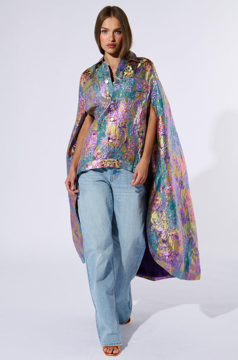 A model walks on a runway wearing the shimmering multicolored VERSAILLES HIGH LOW BROCADE CAPE paired with a matching shirt. The ensemble is completed with loose, light blue jeans and open-toe heels. The plain white background accentuates the vibrant textures and colors of the high-low cape silhouette and shirt. 1