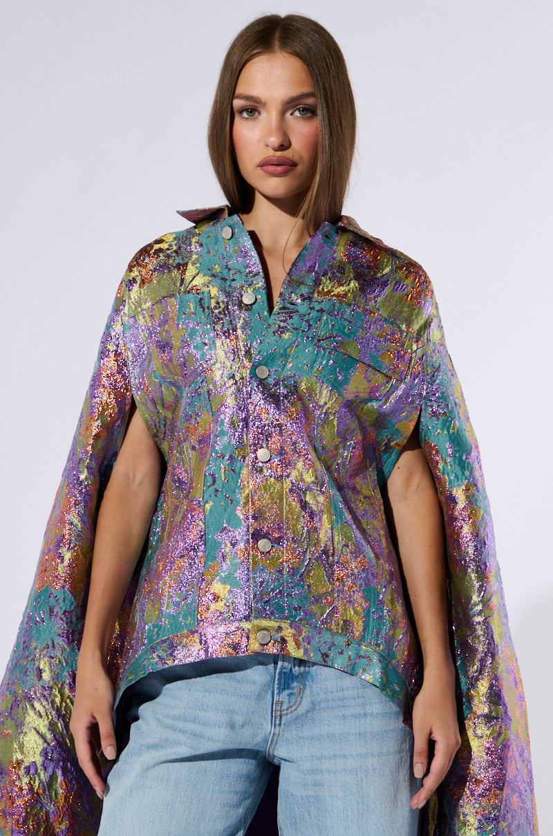 A woman with long brown hair stands against a plain background, showcasing the Versailles High Low Brocade Cape. The shimmery, multicolored metallic brocade cape features rich teal, purple, and gold hues and is styled over a denim shirt and light blue jeans. She has a neutral expression and her hands are relaxed at her sides. 2