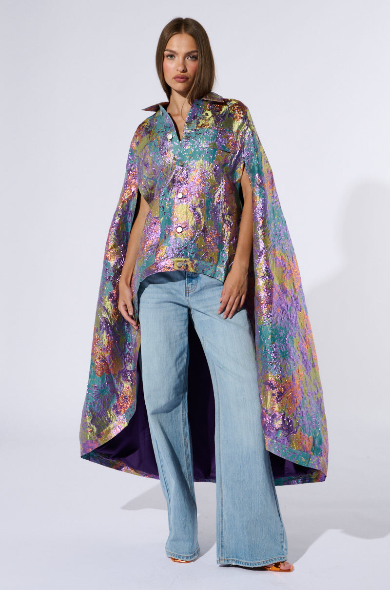 A woman stands confidently against a plain background, wearing the VERSAILLES HIGH LOW BROCADE CAPE, an iridescent piece with a vibrant floral pattern over a matching blouse. She pairs it with wide-legged light blue jeans and orange open-toe heels, her straight shoulder-length hair completing the look. 0