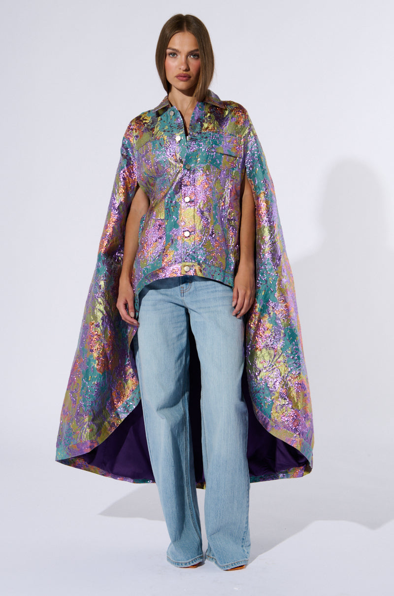 A person stands confidently wearing the VERSAILLES HIGH LOW BROCADE CAPE, a vibrant multicolor metallic brocade piece with a high-low silhouette. They pair it with a matching shirt and light blue, wide-leg jeans. The cape, inspired by Versailles and featuring a purple inner lining, complements their straight, brown hair falling loosely around their shoulders. 3