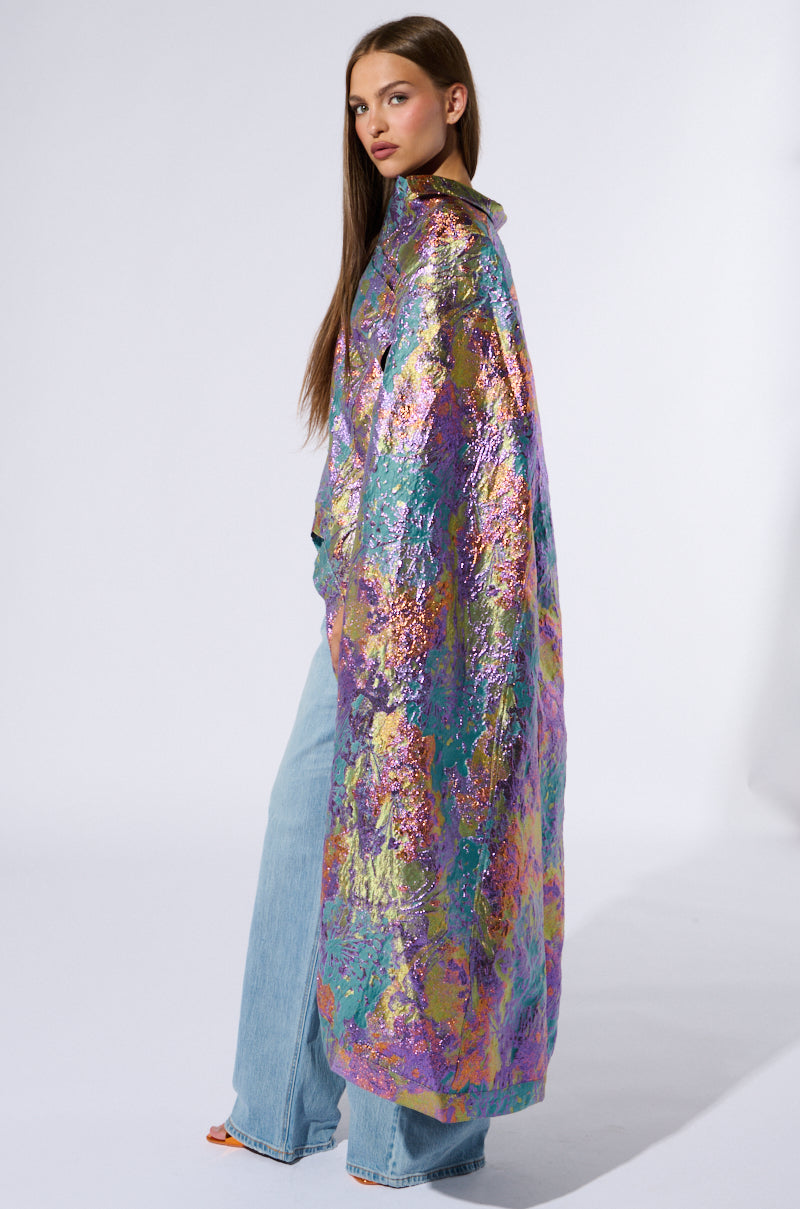 A woman with long, straight hair poses in a colorful, iridescent Versailles High Low Brocade Cape that showcases a vibrant blend of colors and glossy textures. She pairs it with light blue, wide-legged jeans and orange heels against a plain white background. 4