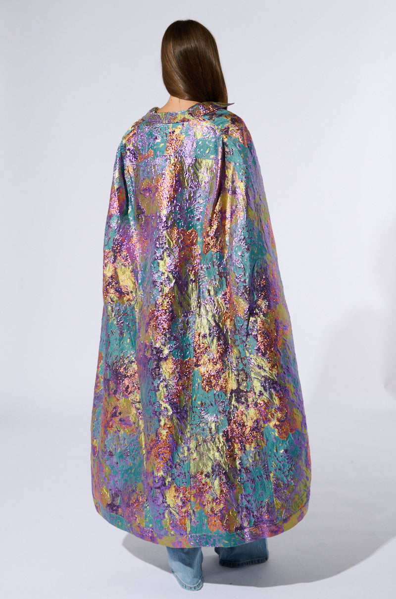 A person with long brown hair is shown from behind wearing the Versailles High Low Brocade Cape, which features a multicolor metallic brocade blend of purple, gold, blue, and green. The high-low silhouette of the cape elegantly drapes down to their ankles as they stand against a plain, light-colored background. 5