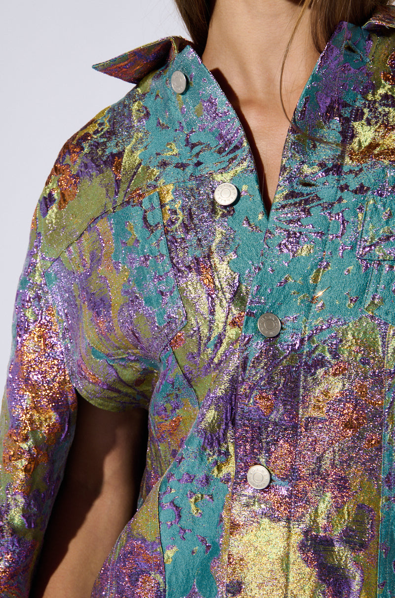 Close-up of a person wearing the Versailles High Low Brocade Cape. The fabric features a vibrant, multicolored abstract pattern with shades of purple, green, gold, and turquoise, adorned with shiny silver buttons. The material has a textured, shimmering appearance reminiscent of multicolor metallic brocade. 6