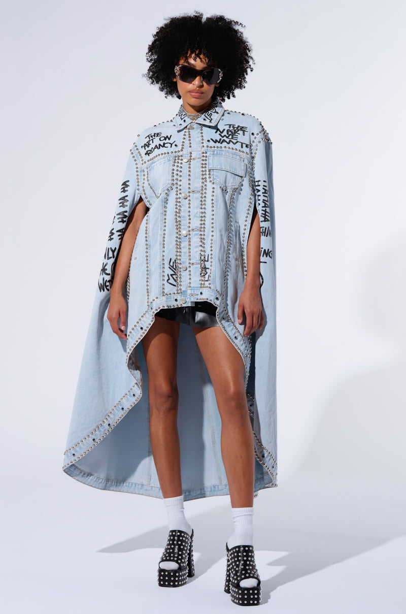 A person stands against a light background wearing the RHYTHM OF THE NIGHT DENIM CAPE, a light blue, asymmetrical denim top embellished with metallic silver studs and black graffiti patterns. They are paired with dark shorts, platform sandals with white socks, and sunglasses. The ensemble includes a cape-like flow at the back.