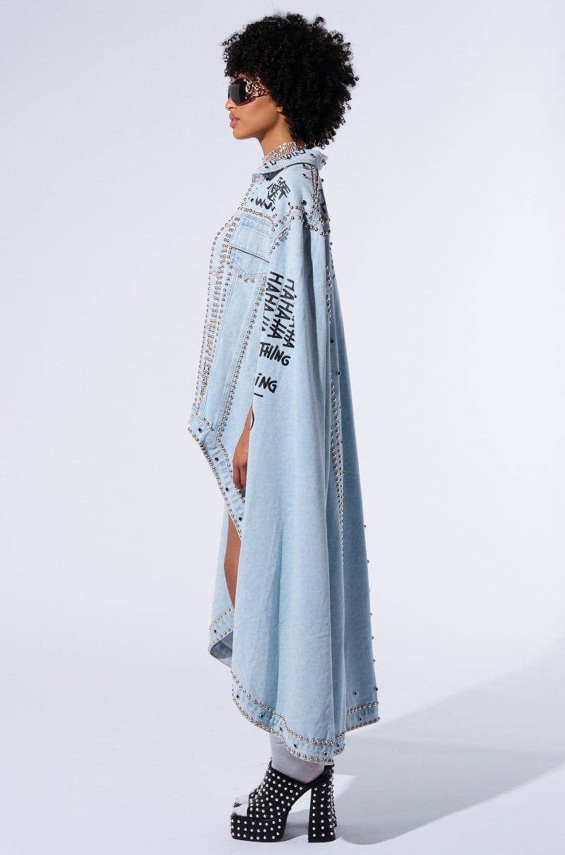 A person with curly hair wearing dark sunglasses and a long, light RHYTHM OF THE NIGHT DENIM CAPE adorned with metallic silver stud embellishments poses in profile. The cape features street-inspired graffiti on the back and sides, and the individual is also wearing platform shoes with studded details.