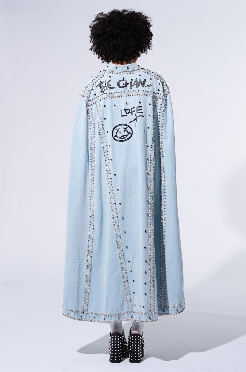 A person with curly hair, seen from the back, is sporting the RHYTHM OF THE NIGHT DENIM CAPE, a long and light blue garment decorated with metallic silver stud embellishments and graffiti-style writing that includes "THE CHAIN" and "LOVE." They pair it with black studded boots against a plain, light-colored background.
