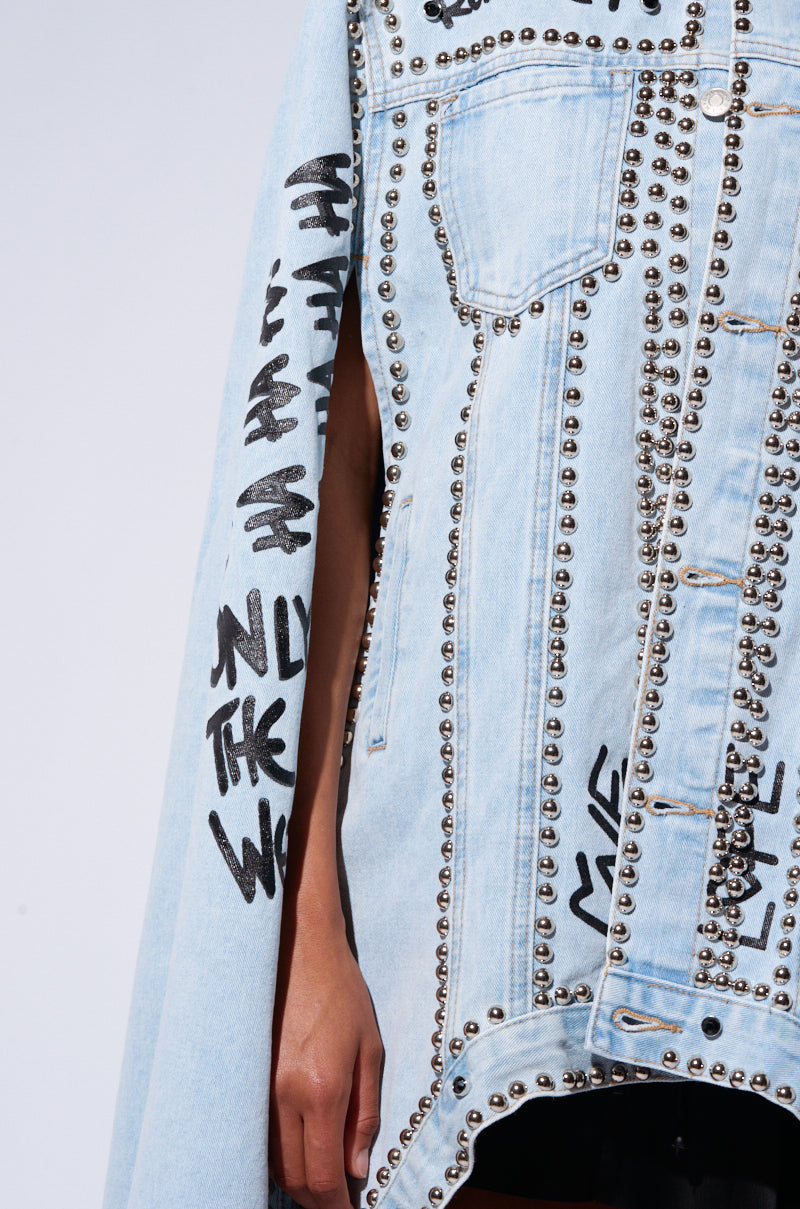 Close-up of a person wearing the RHYTHM OF THE NIGHT DENIM CAPE, a light blue denim jacket adorned with metallic silver stud embellishments arranged in patterns along the seams and edges. The cape features street-inspired graffiti-like text and designs, with a partial view of the person's arm visible through an open side.